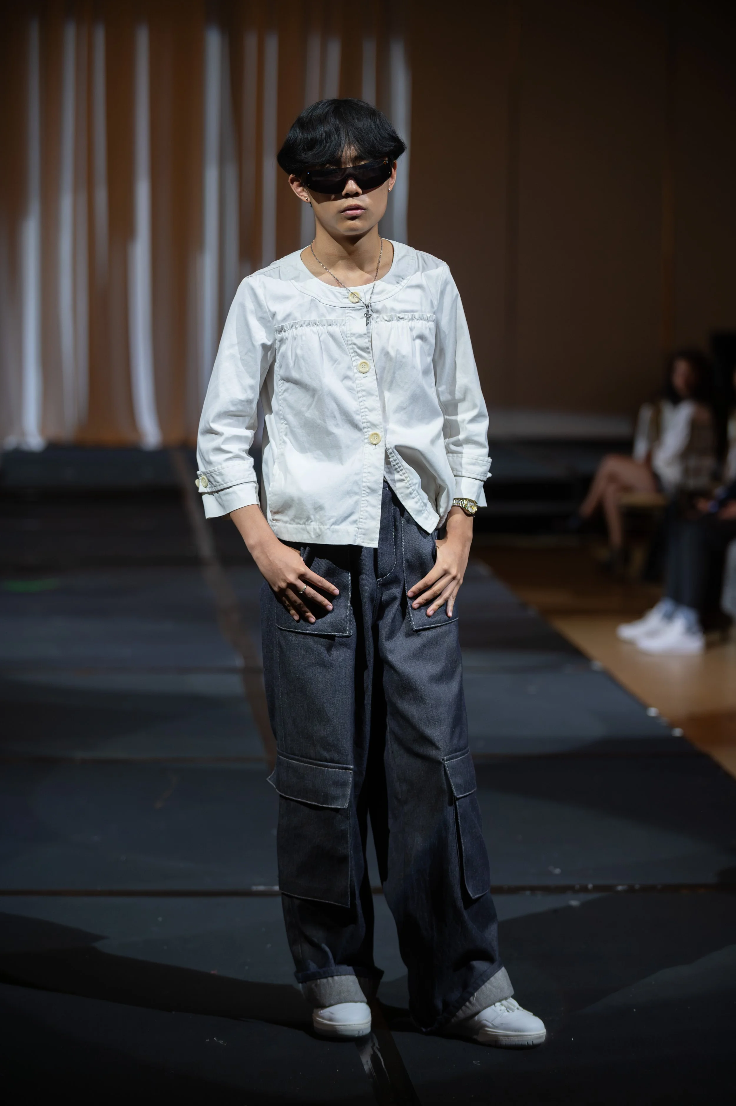 A young person with black hair wearing sunglasses, a white button-up shirt, baggy dark jeans, white shoes, and jewelry, standing on a stage with curtain backdrop and another person seated in the background.