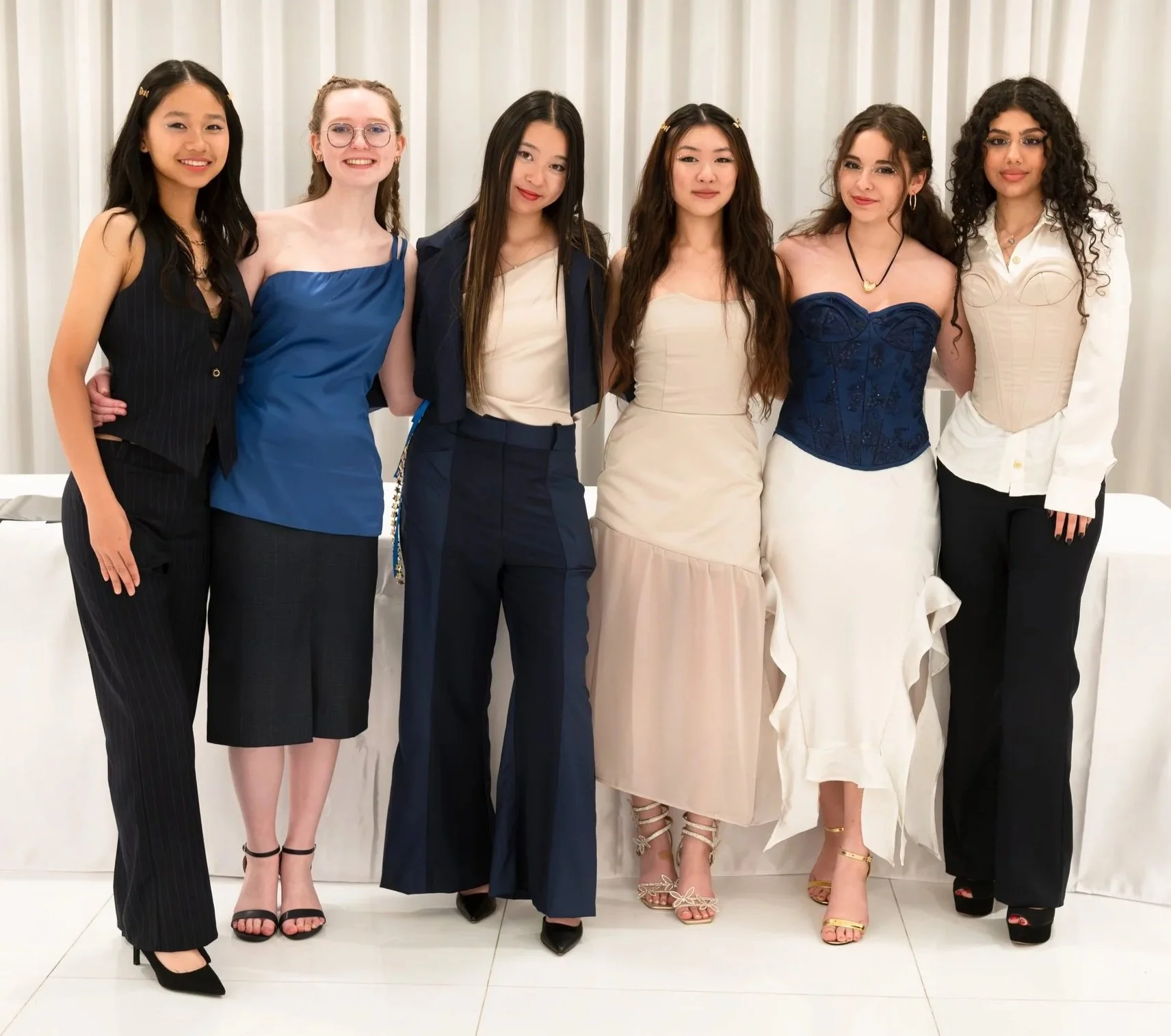 Group of seven women dressed in formal attire, standing together in front of a curtain. They are smiling and have their arms around each other.