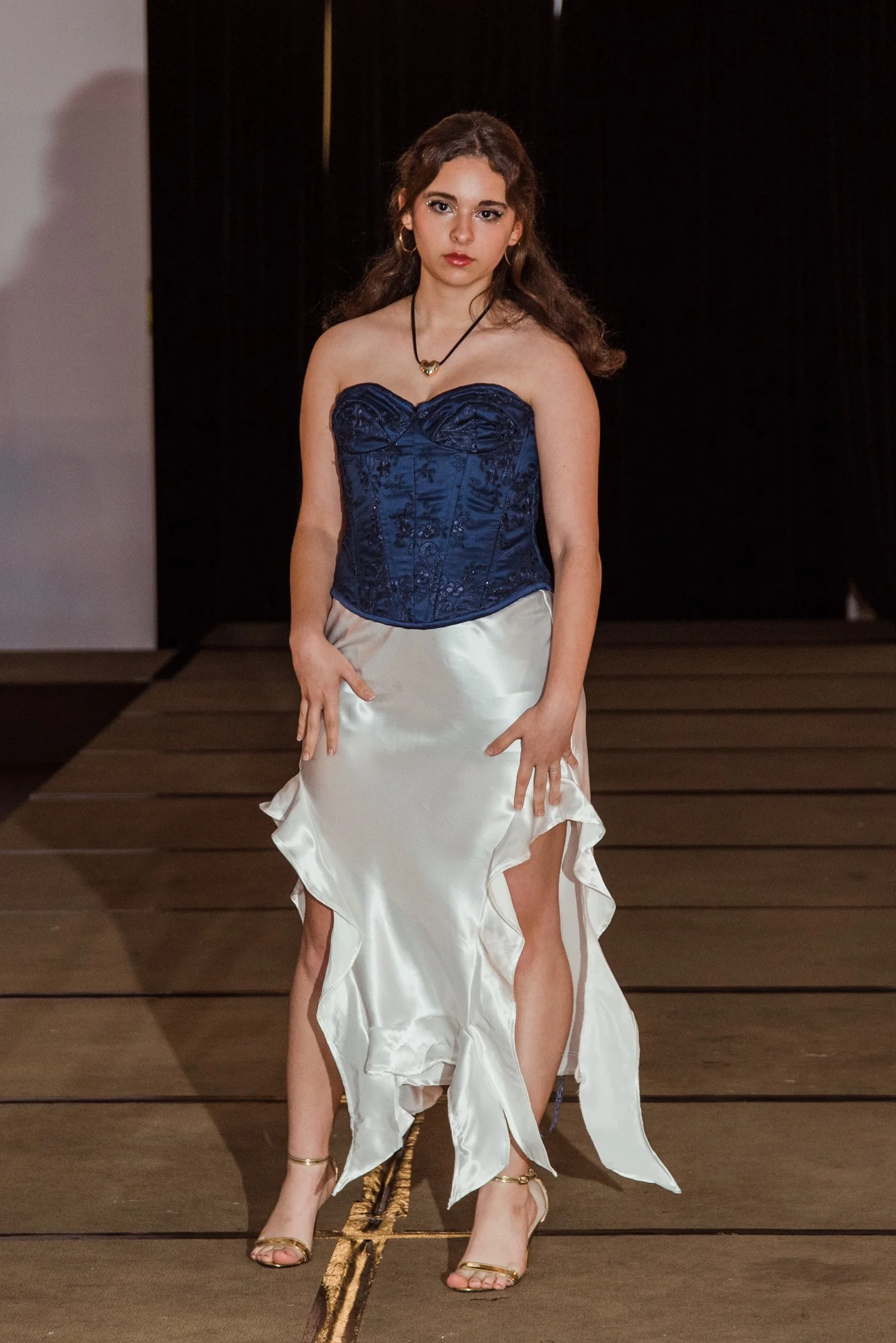 A young woman with long, curly brown hair and light skin wearing a strapless blue corset and a white satin skirt, standing on a runway. She is accessorized with a black necklace with a gold heart pendant, gold hoop earrings, and gold heels, with a dark background behind her.