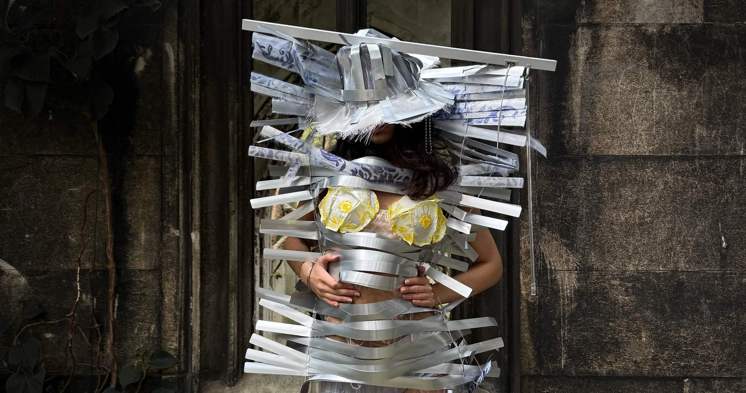 Model wearing garment and hat made out of aluminium window blinds, with yellow floral bra top peaking from under.