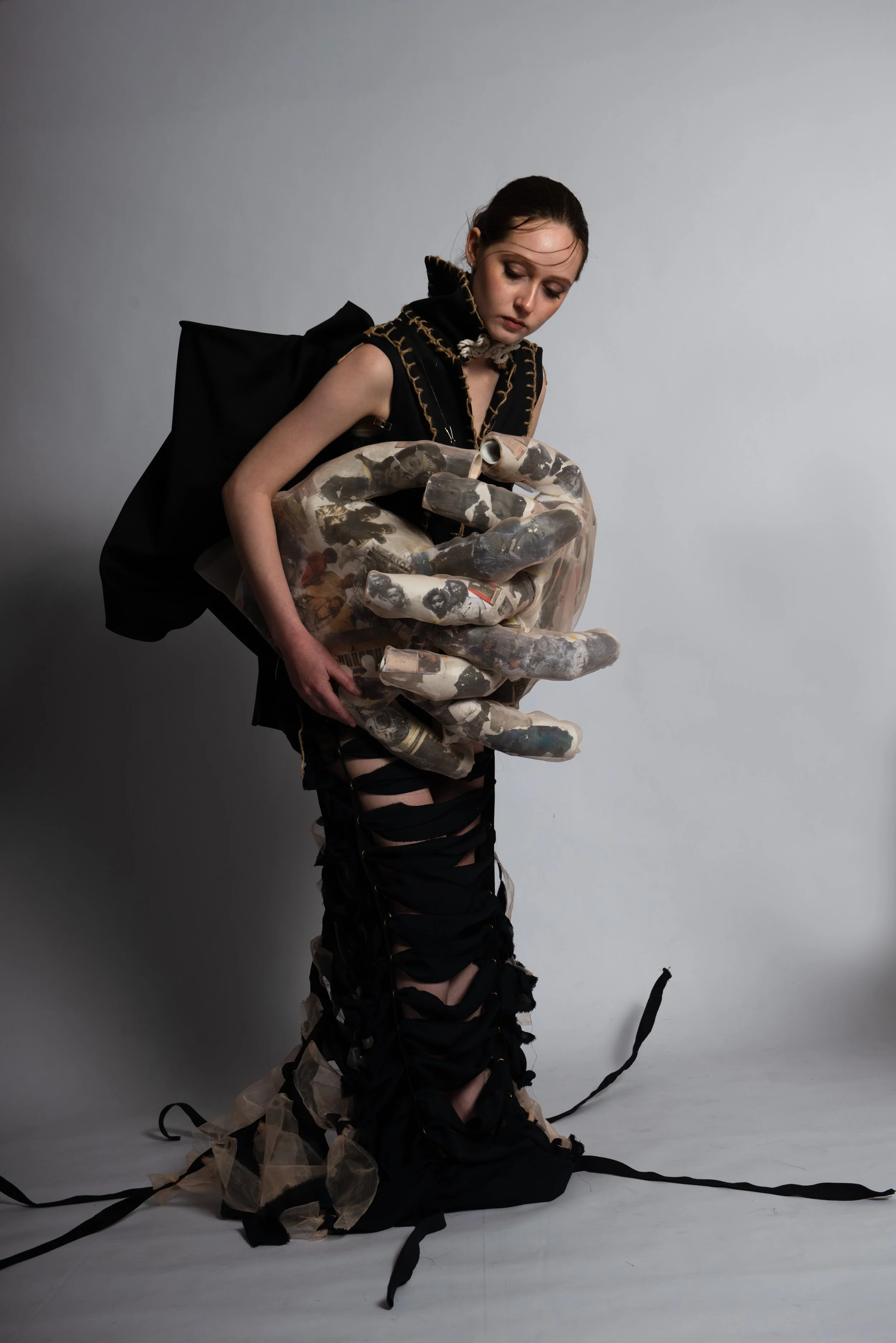 A woman with dark hair styled back, wearing a black outfit with gold-stitch detailing, holds a large, sculptural, hand-shaped object made of collage-like material featuring black and white images, standing against a plain light gray background.