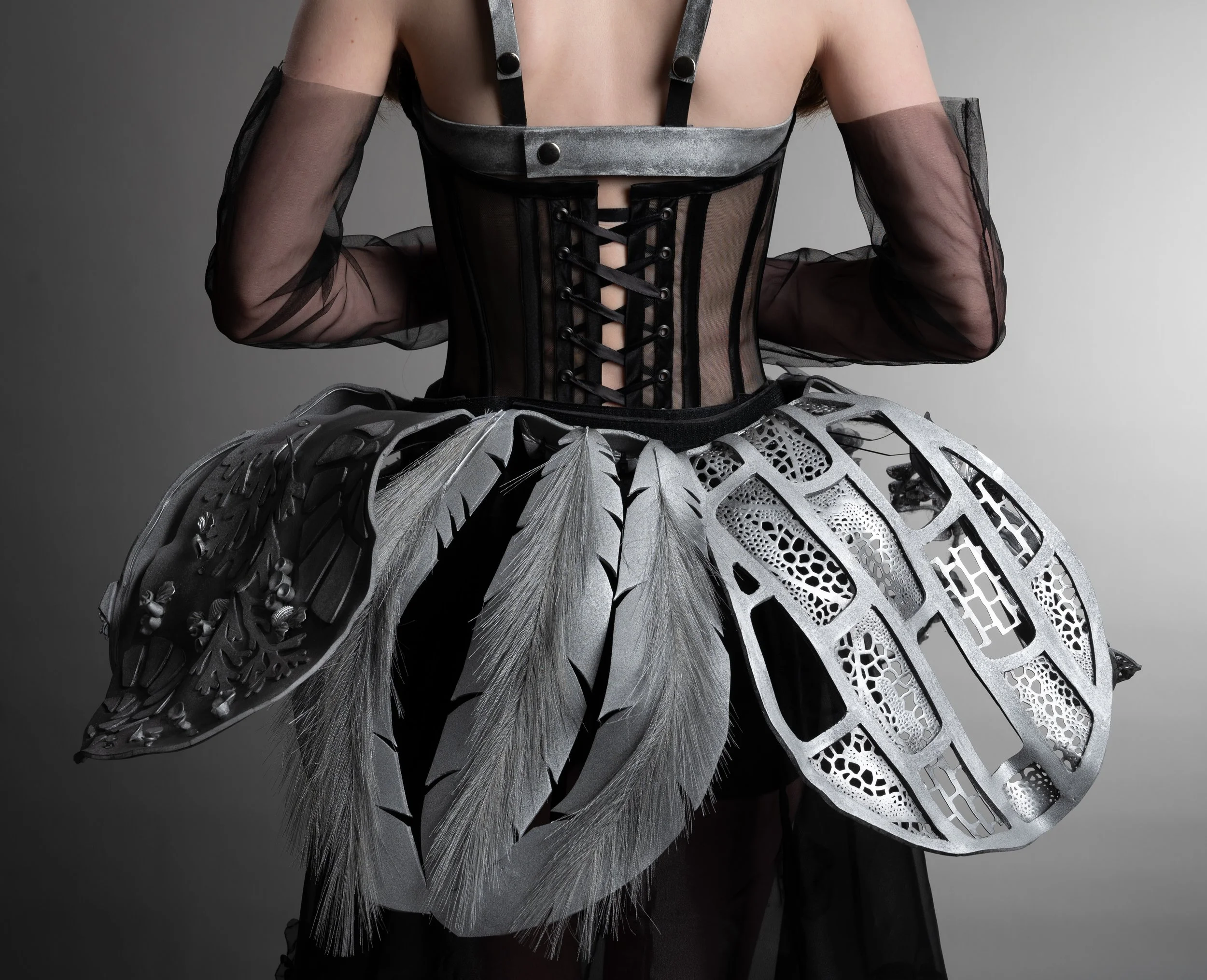 Close-up of a person wearing a black sheer top with puffed sleeves and a metallic corset with lacing detail over it. The person is also wearing a futuristic, metallic, leaf-like skirt or accessory with intricate cut-out patterns.