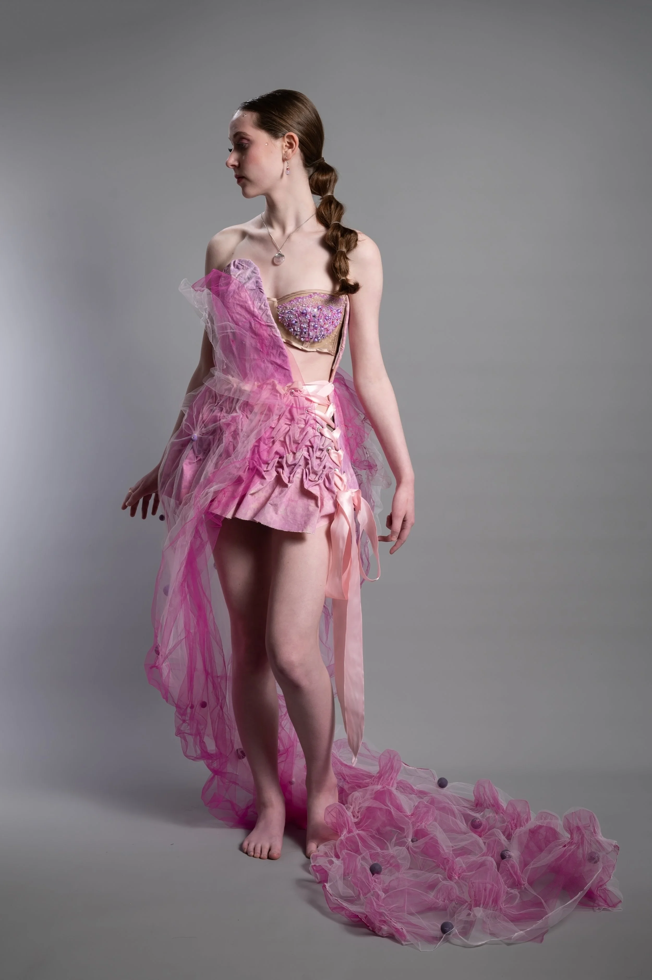 A young woman with long, brown hair styled in a braid. She is wearing a pink, sheer, ruffled gown with a fitted bodice adorned with colorful beads and sequins. She is standing on a plain gray background, looking down and to her left, with one arm slightly extended and the other relaxed at her side. A pink tulle fabric with balls is draped around her, trailing on the floor.
