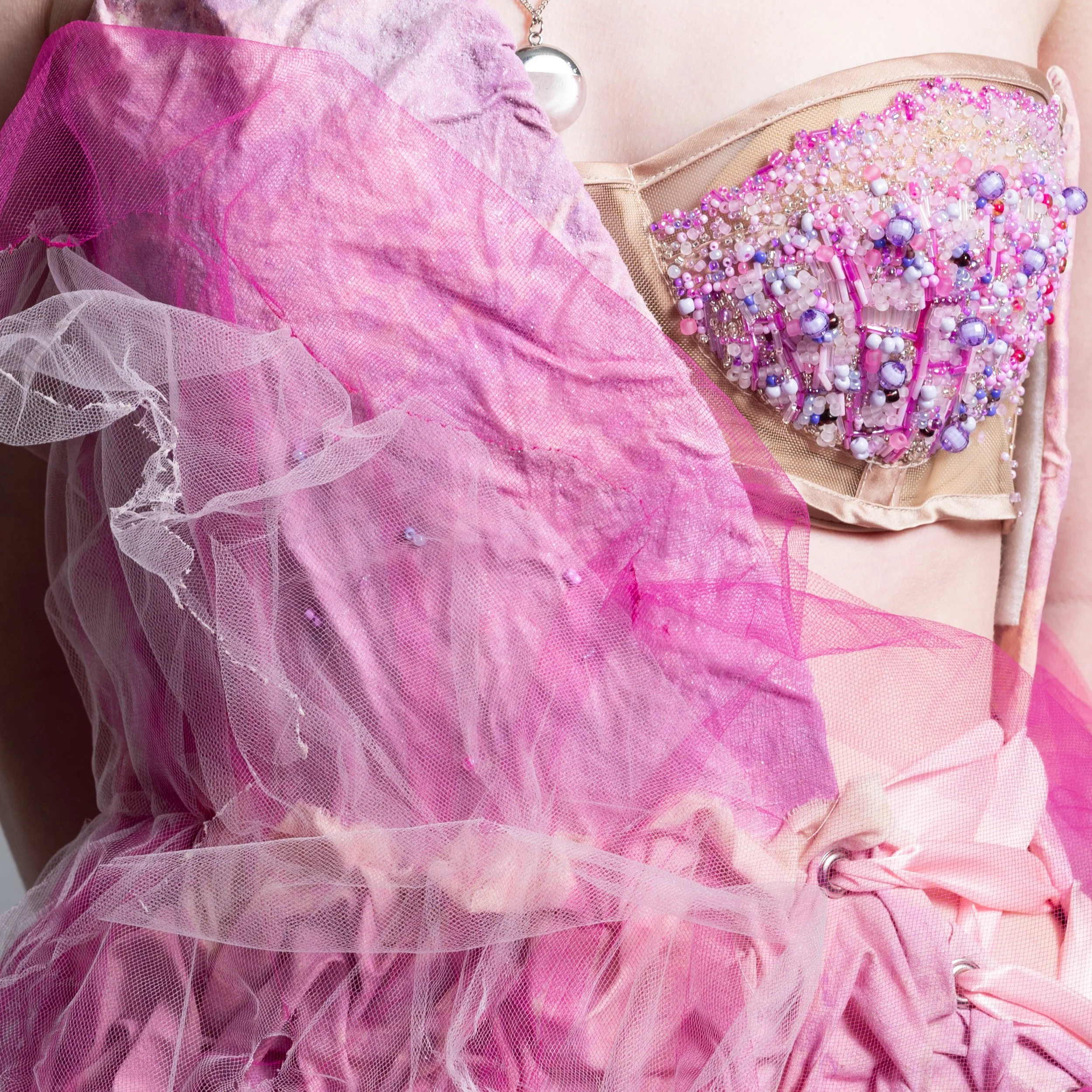 Close-up of pink tulle with beads and rhinestones, part of a decorative accessory or costume, with other pink fabric and a large pearl necklace in the background.