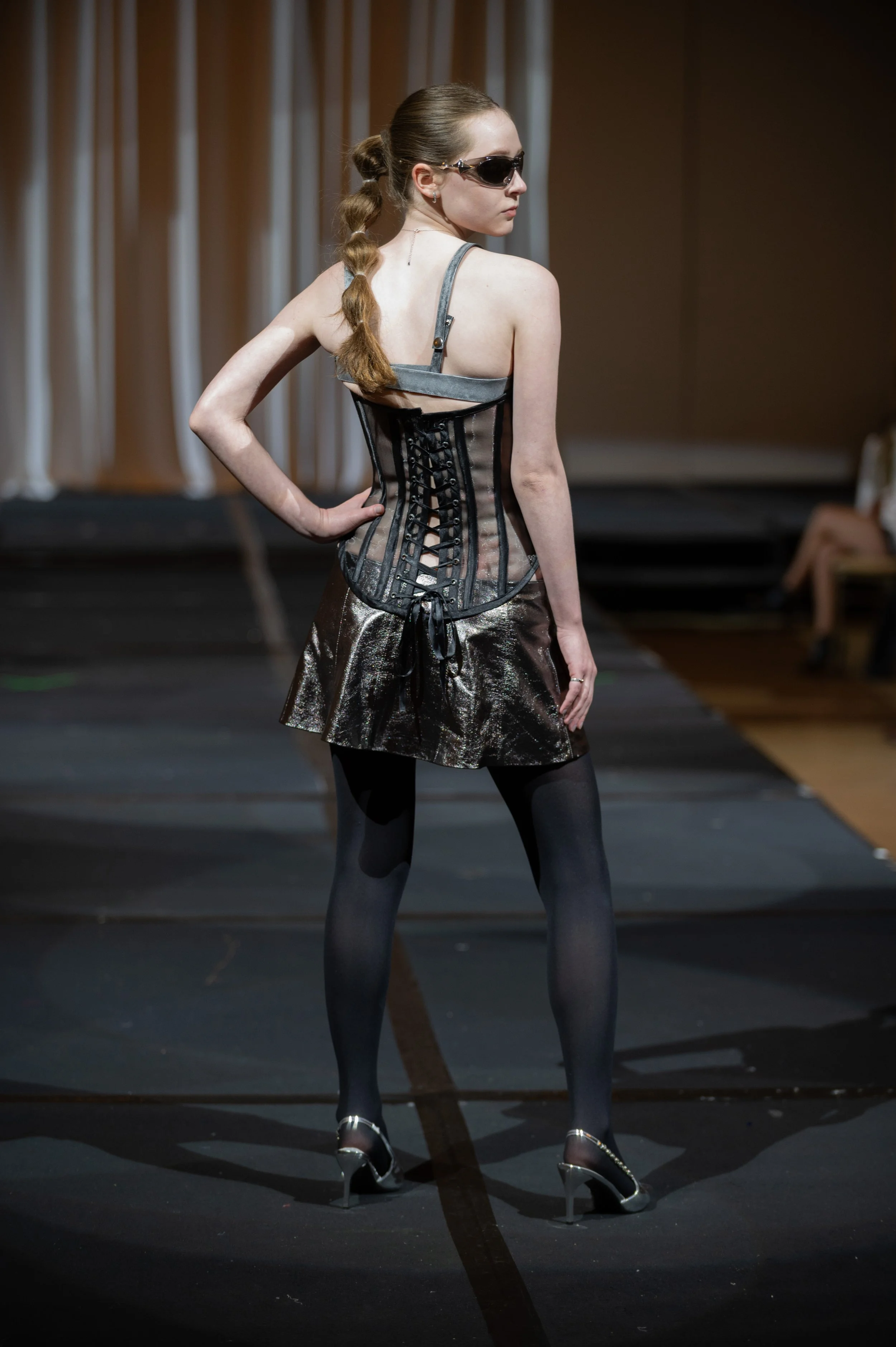 A woman on a runway model wearing sunglasses, a grey corset top, metallic skirt, and black tights with high heels, posing with her back partially turned.