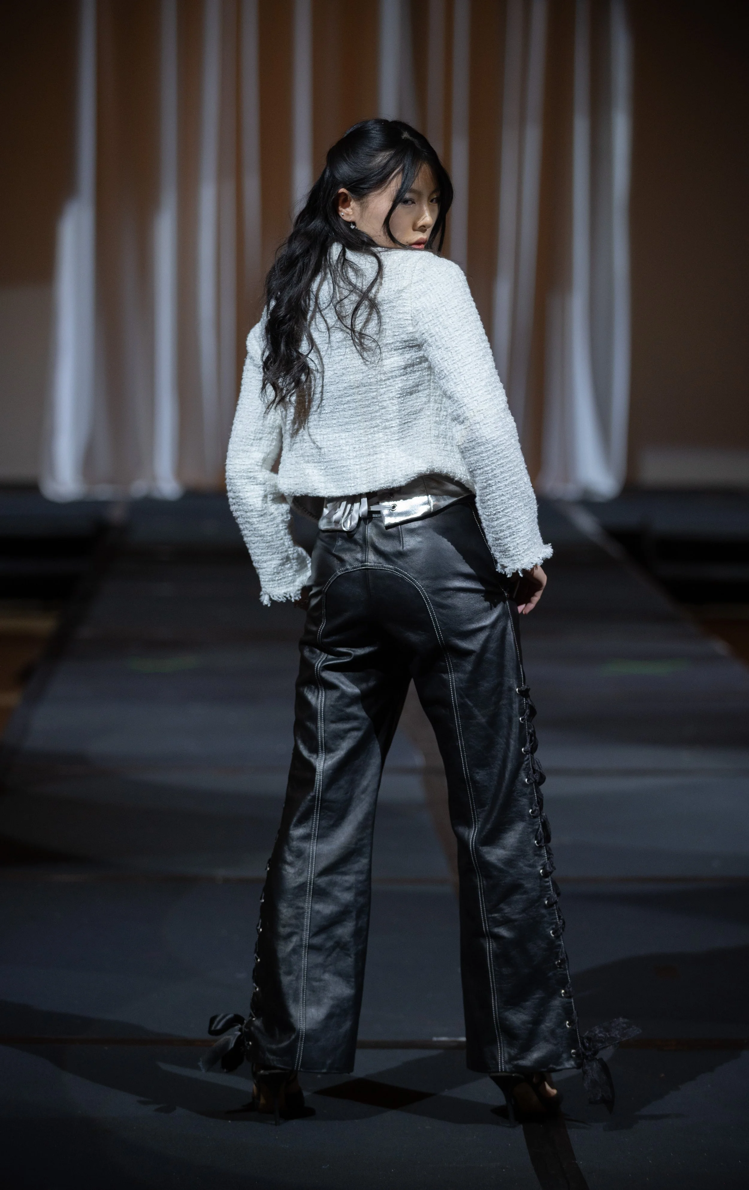 A woman with long black hair in a white textured jacket and black shiny pants walking on a runway with a dark background and curtains.