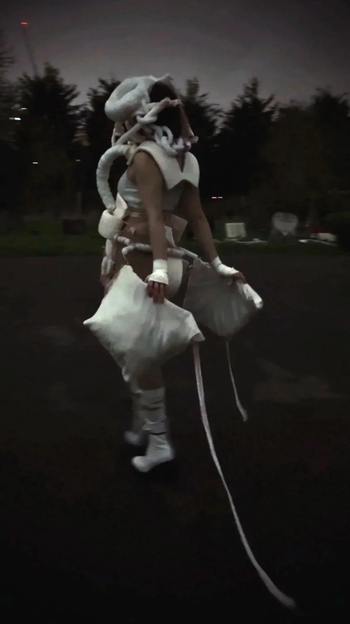 A person wearing white avant-garde clothing, including high-heeled boots, gloves,, standing outside at dusk with trees and park benches in the background.
