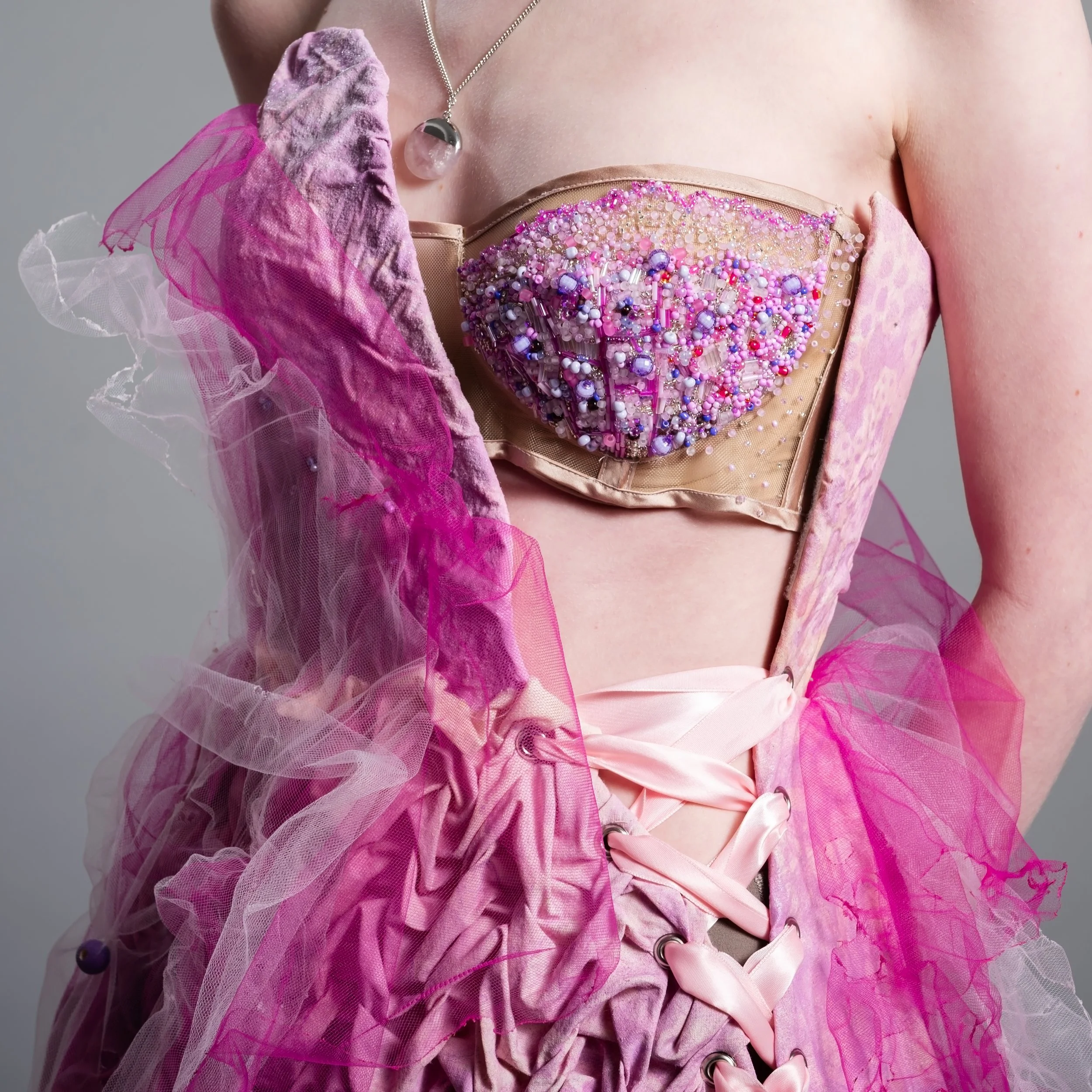 Close-up of a woman wearing a pink, bejeweled, avant-garde corset dress with pink tulle fabric, and a pink satin ribbon at the waist.