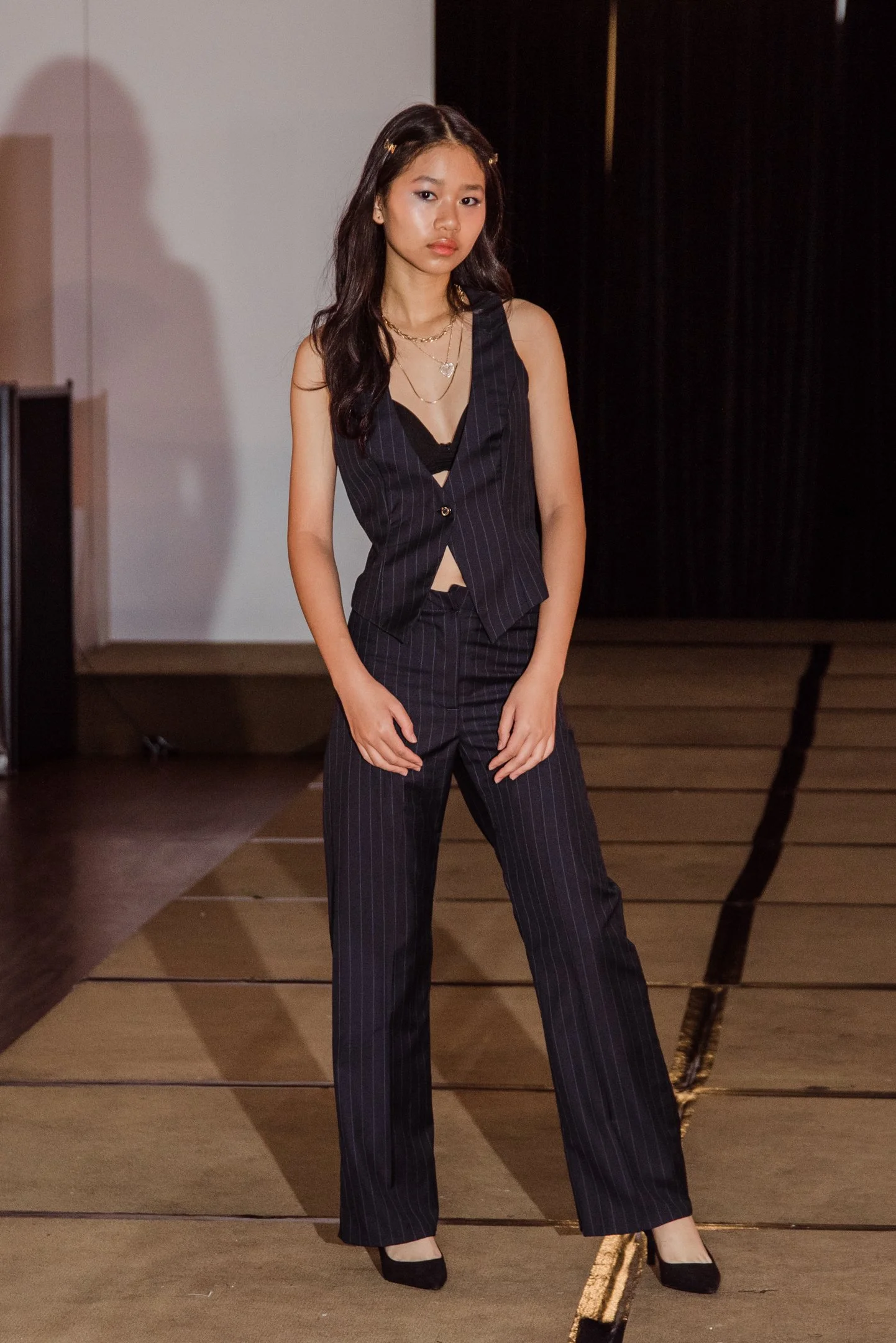 Young woman standing indoors on a stage, wearing a pinstripe suit with a sleeveless vest and wide-leg trousers, accessorized with layered necklaces and black heels, with a black curtain and hardwood floor in the background.