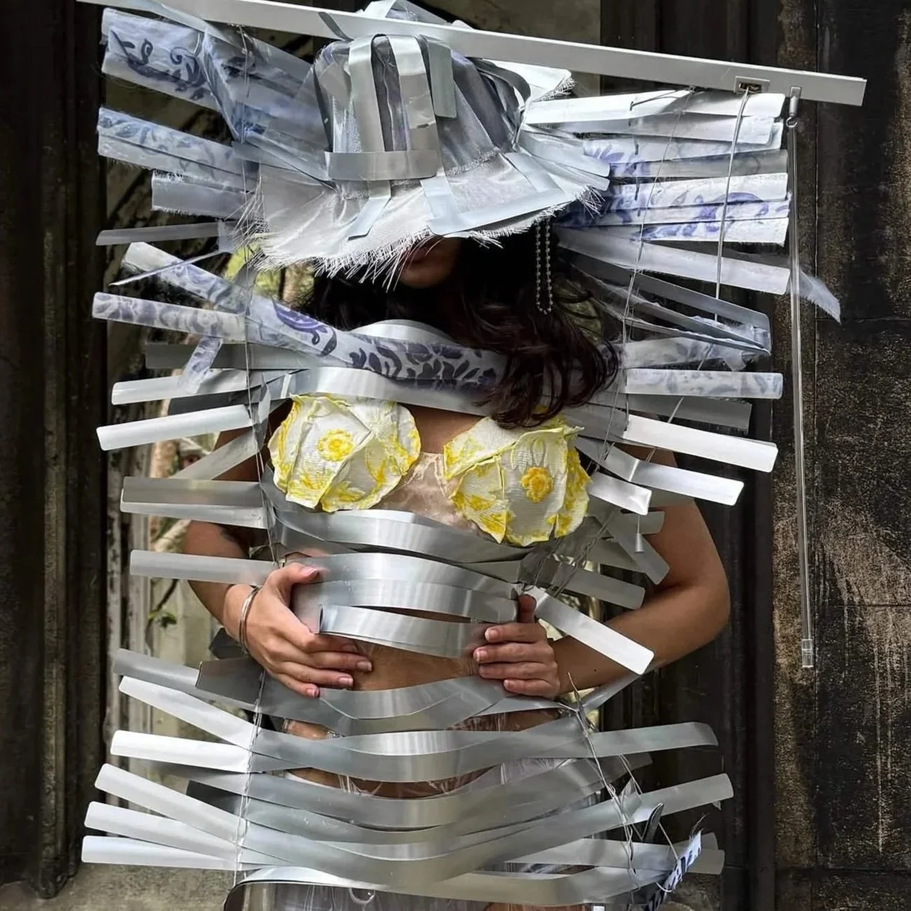 Person standing behind a metallic art installation resembling a face with large hat. The person wears a yellow floral top and holds a concrete object. The installation is made of metallic strips and reflective surfaces.