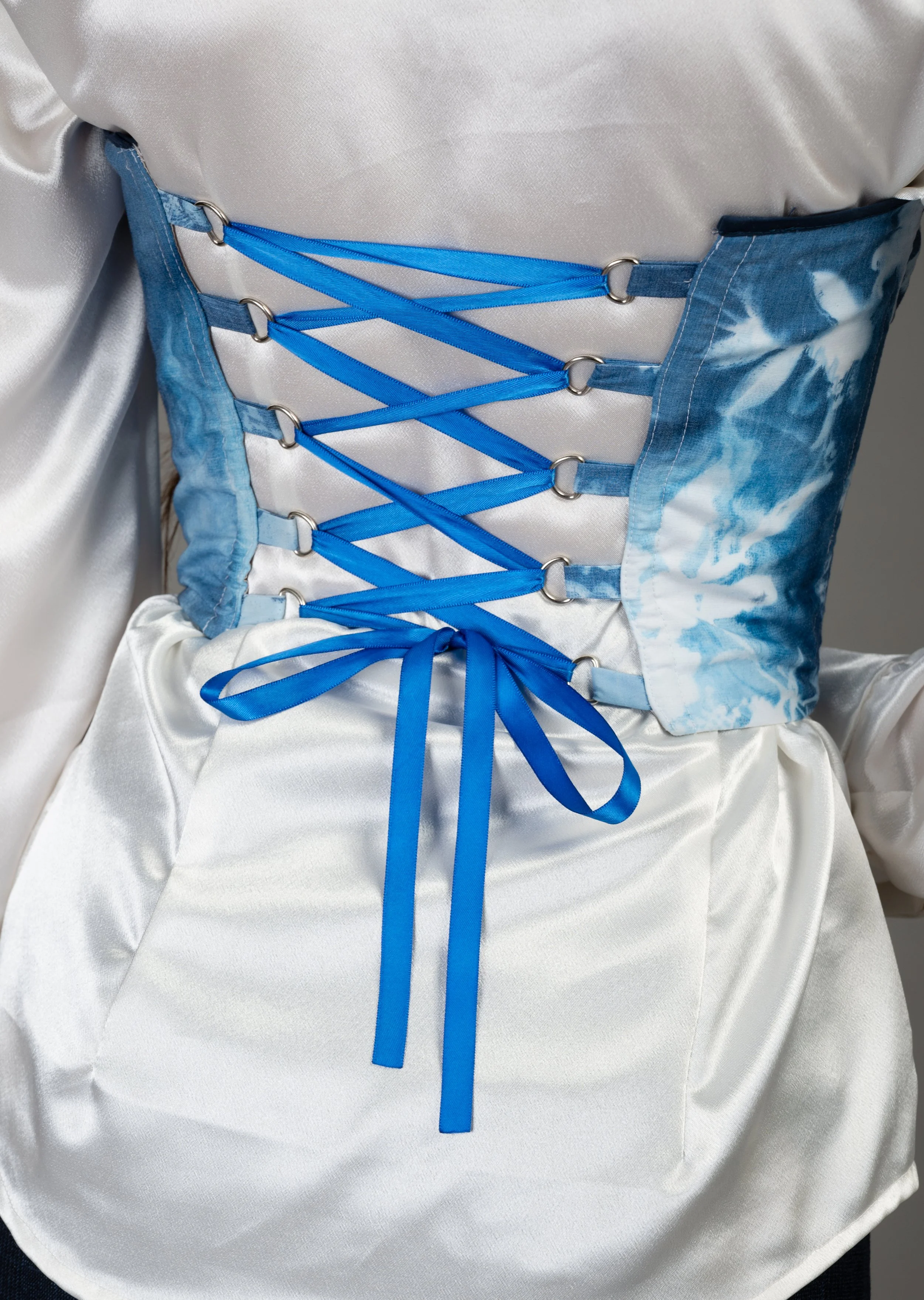 Close-up of a person's waist showing a corset-style lace-up detail with blue ribbons on a shiny, satin fabric background.