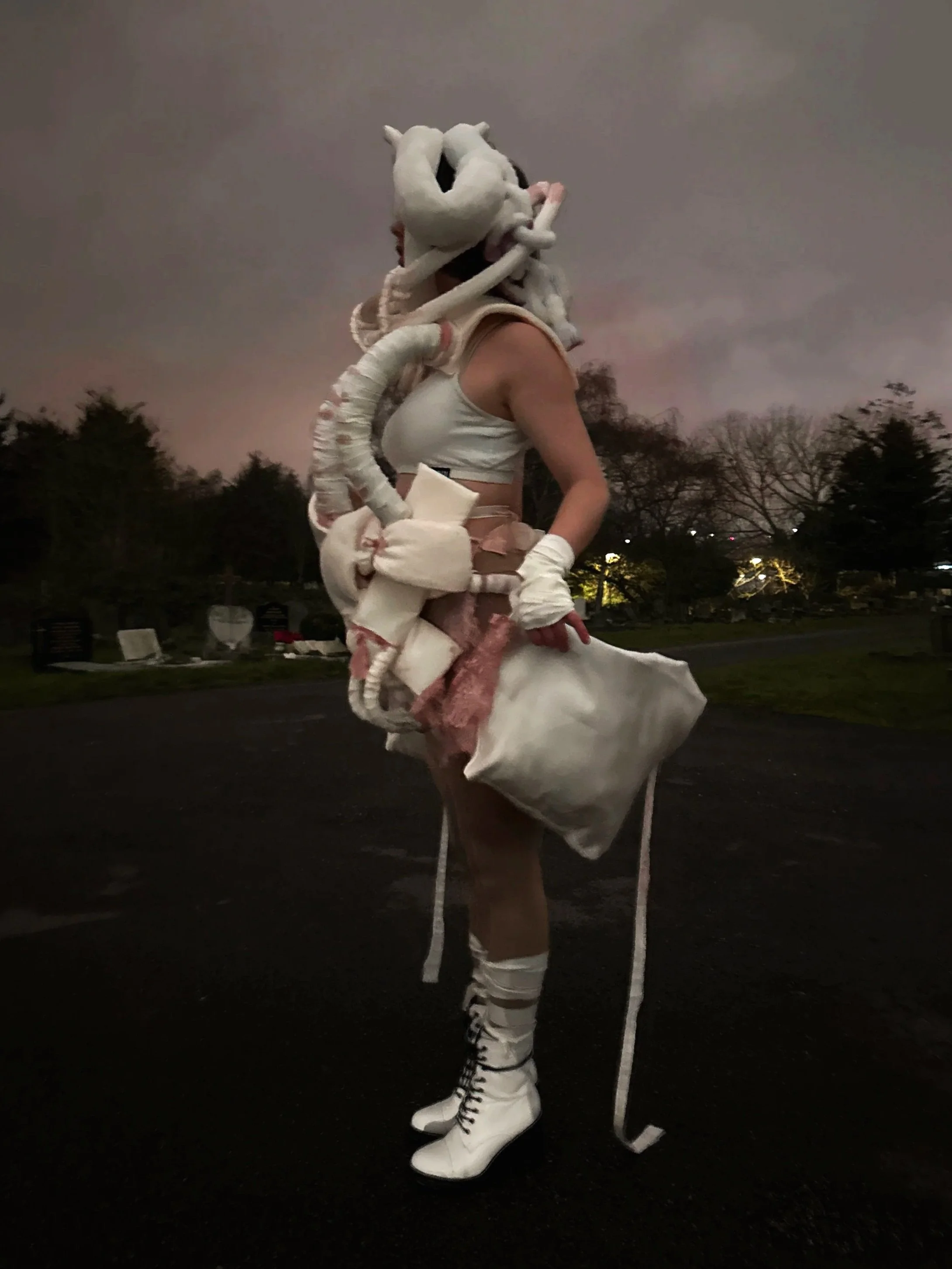 A person wearing white futuristic clothing, including high-heeled boots, gloves, and a skirt, standing outside at dusk with trees and park benches in the background.