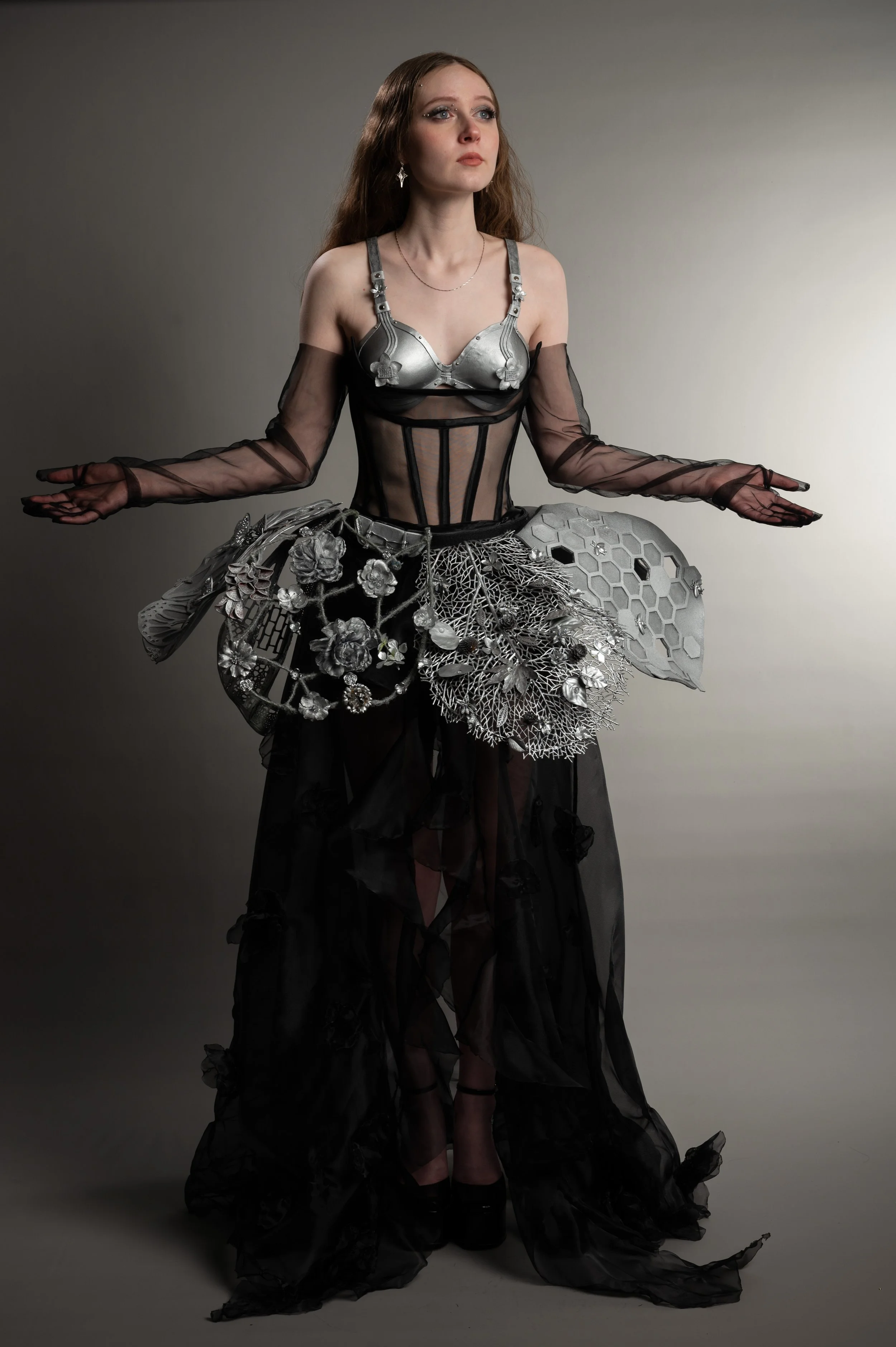 A woman with long red hair wearing a metallic silver bra, black sheer fabric top with long sleeves, and a black skirt with intricate silver floral embellishments, posing against a neutral background.