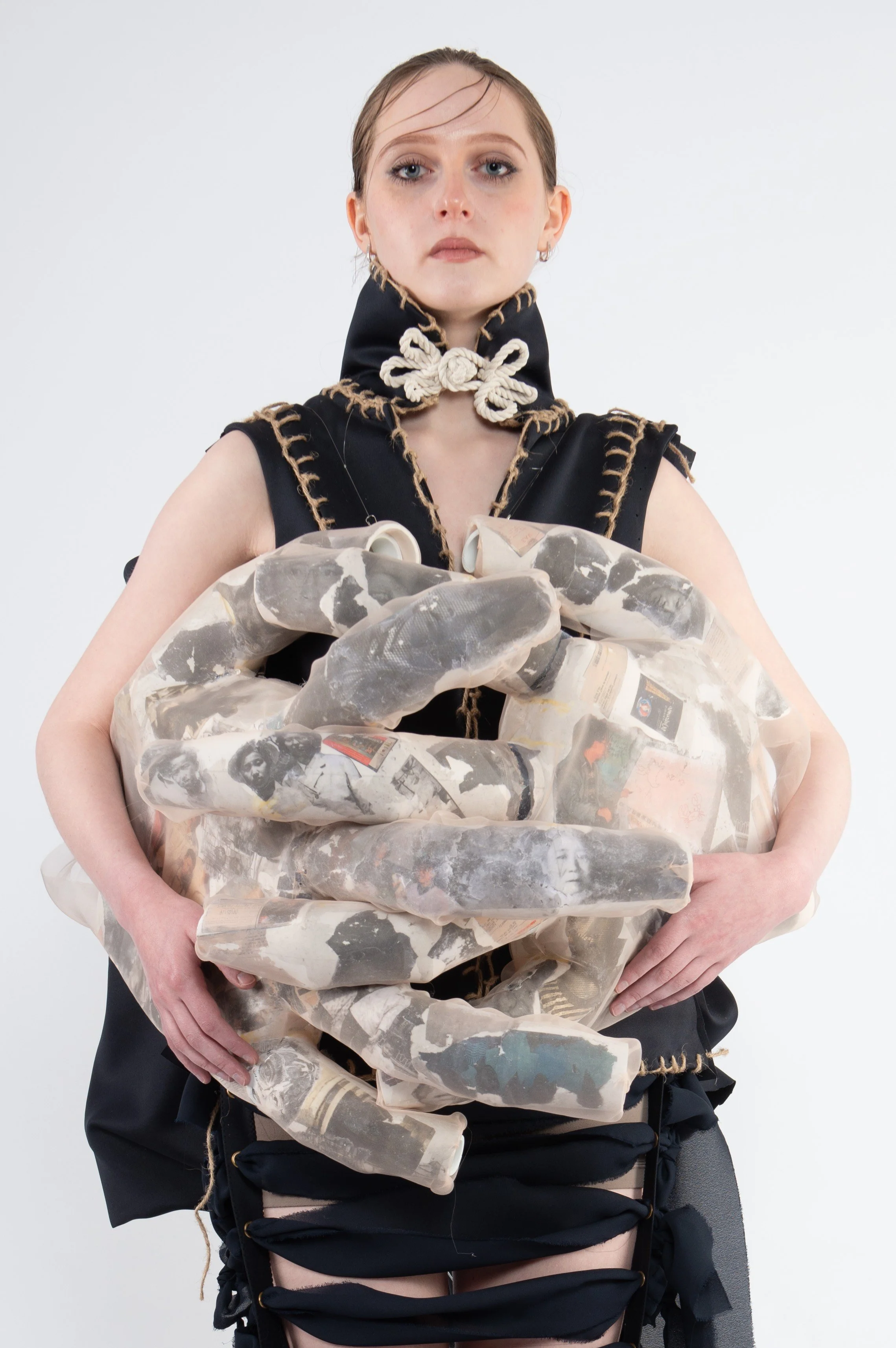 A woman with dark hair styled back, wearing a black outfit with gold-stitch detailing, holds a large, sculptural, hand-shaped object made of collage-like material featuring black and white images, standing against a plain light gray background.