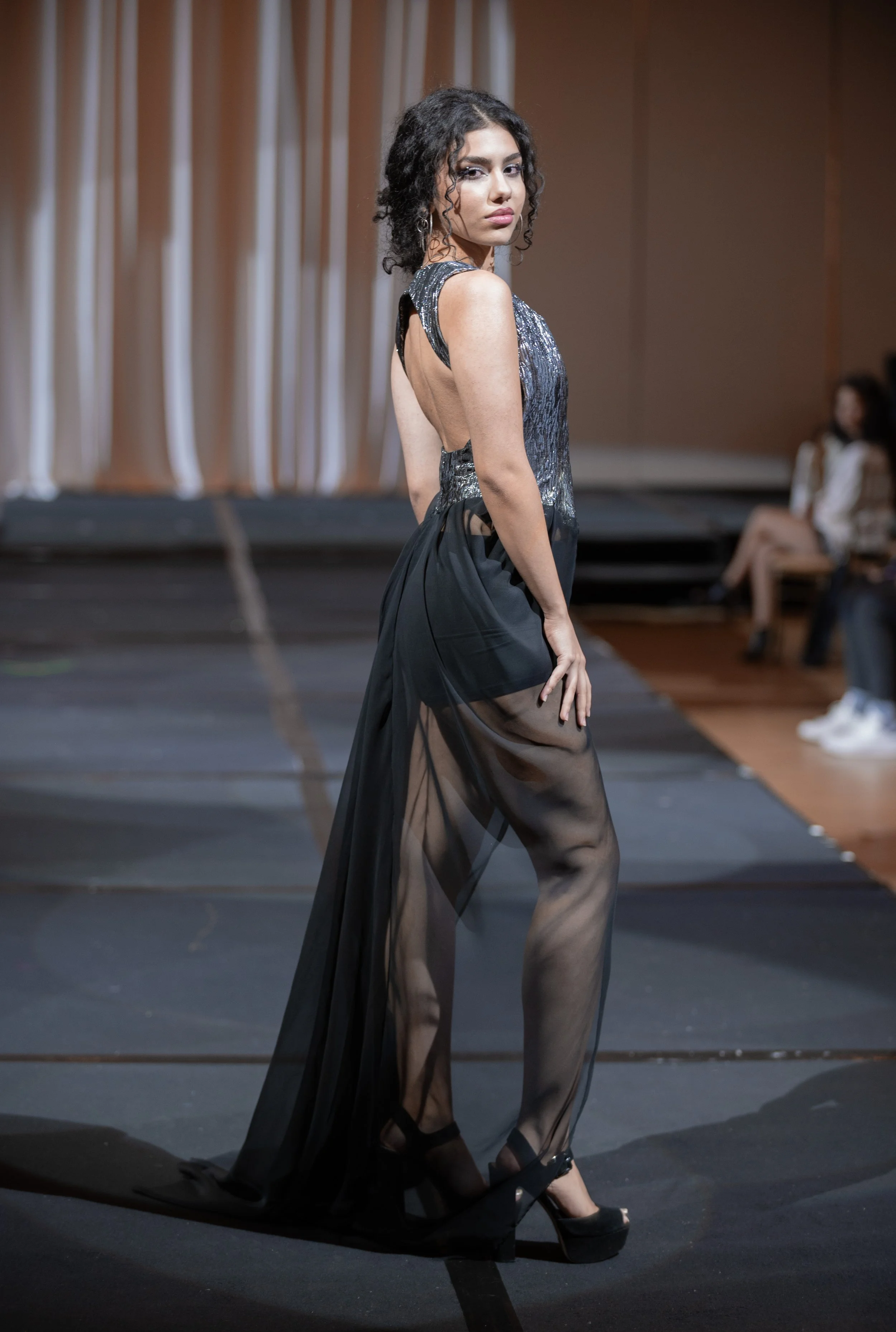 A young woman with curly hair walking on a runway in a black sheer dress with a glittery top, black high heels, and earrings, with onlookers sitting in the background.
