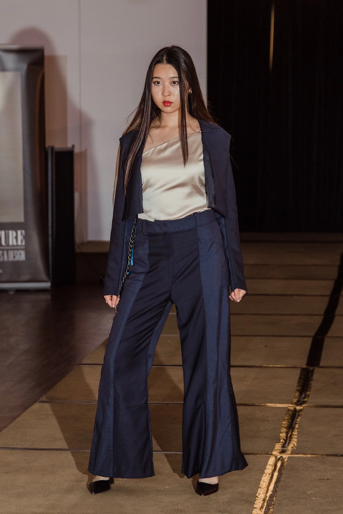 A young woman with long dark hair and red lipstick, wearing a champagne-colored satin top, navy blazer, and wide-leg navy pants, standing indoors near a black curtain.