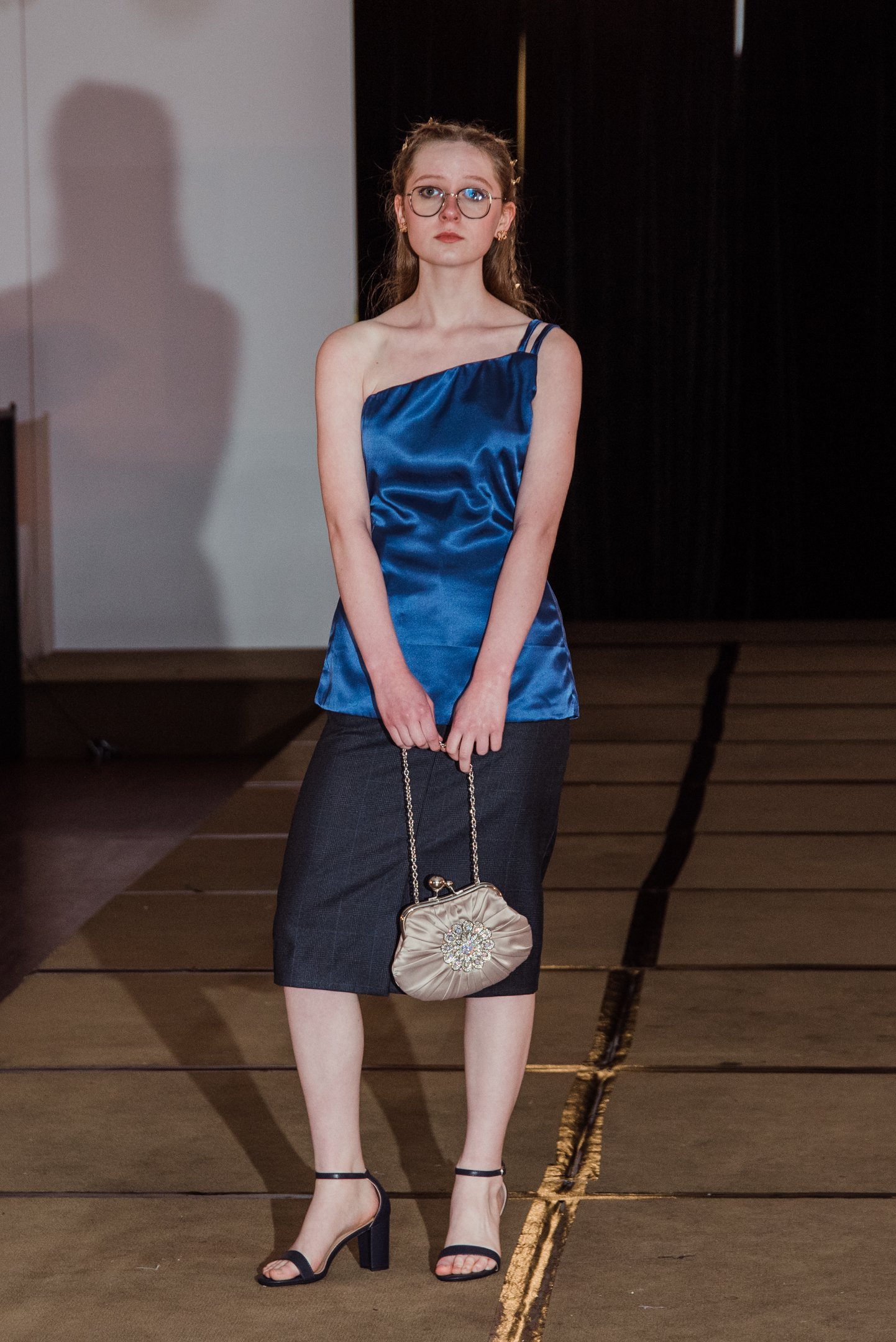 A woman with long red hair, glasses, and earrings wearing a satin blue top, dark gray skirt, black high heels, and holding a small beige purse standing on a runway.
