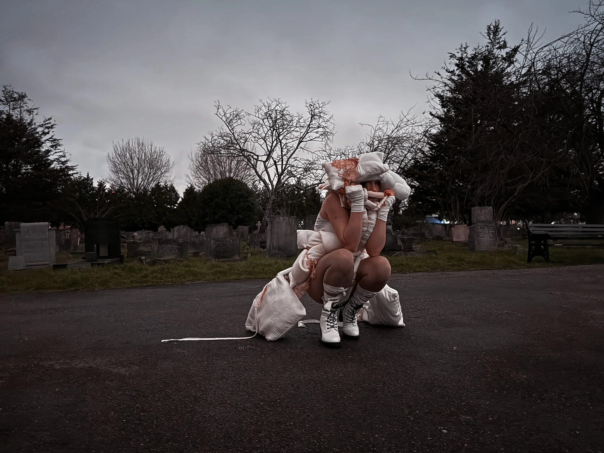A person wearing white avant-garde clothing, including high-heeled boots, gloves,, standing outside at dusk with trees and park benches in the background.