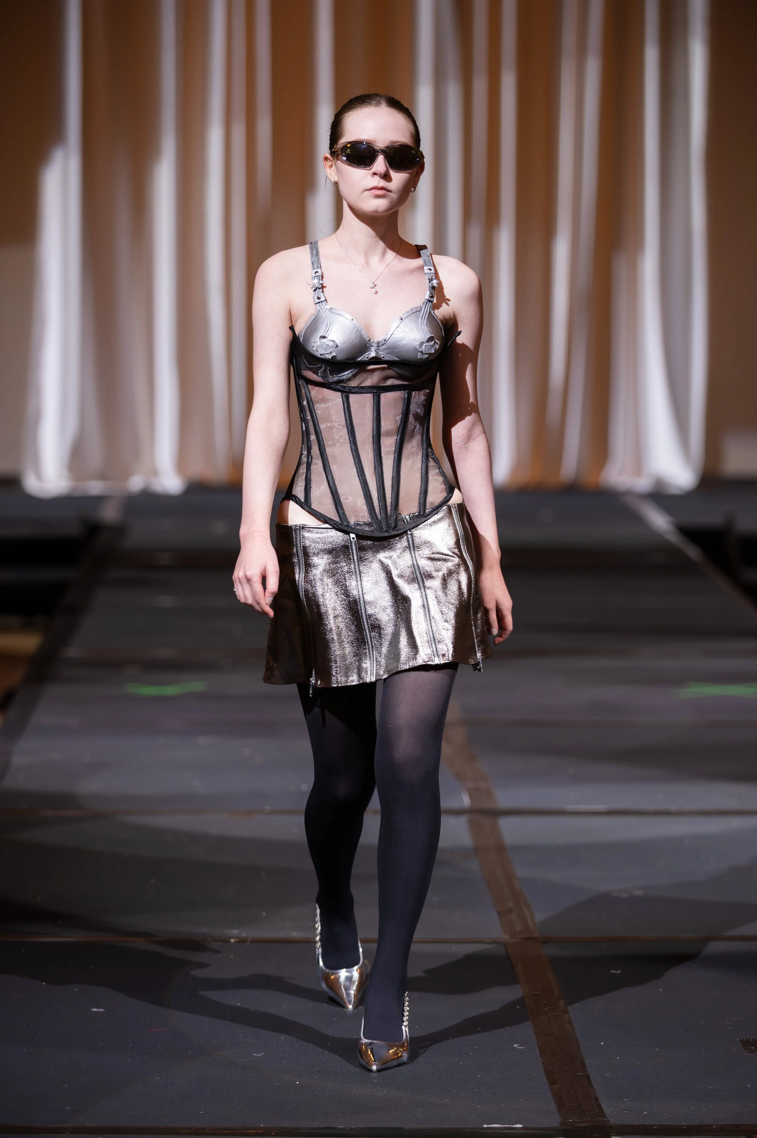 A female model walking on a runway during a fashion show, wearing sunglasses, a metallic bra top, a sheer corset, metallic skirt, black tights, and shiny high heels.