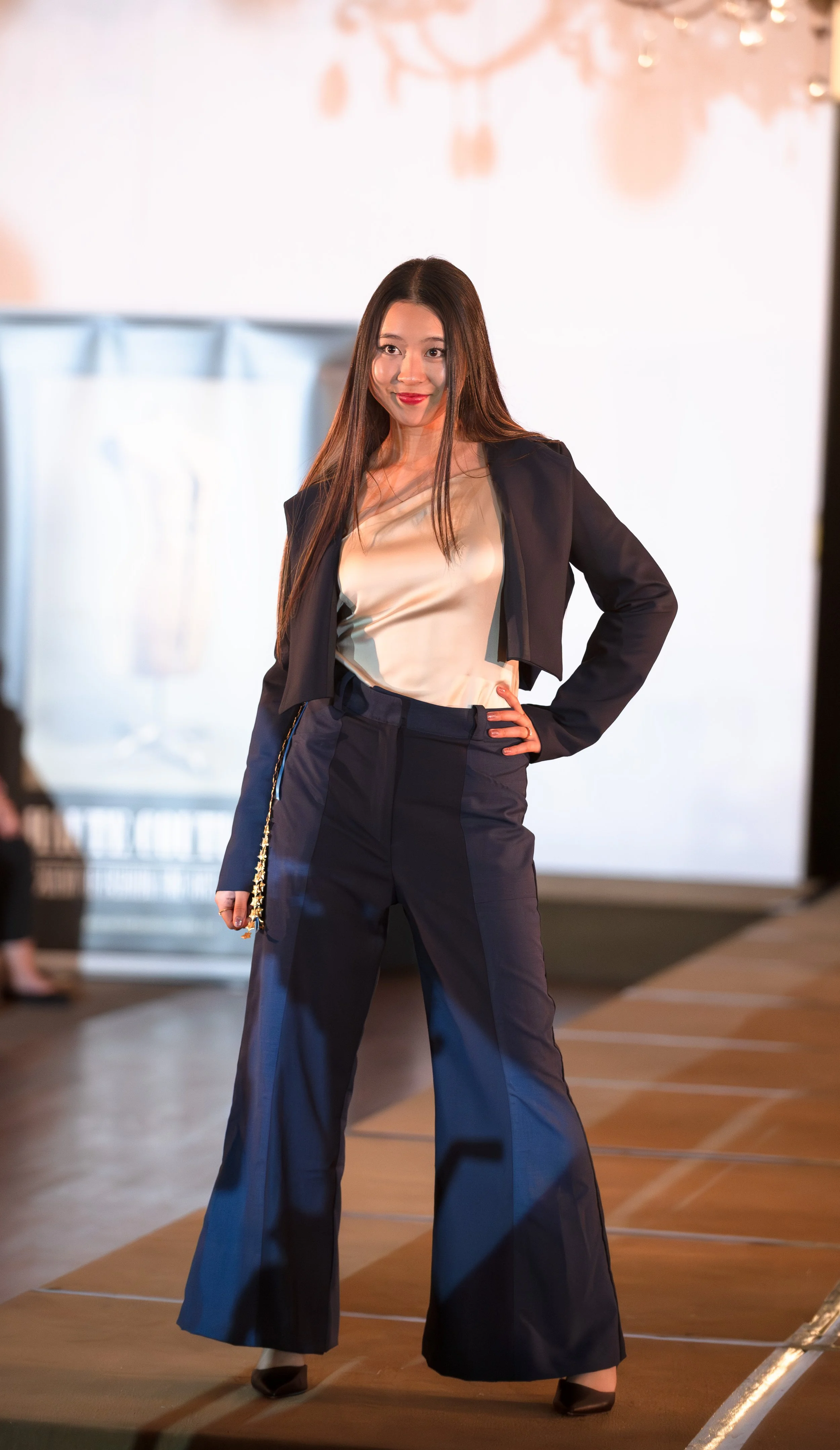 A woman with long brown hair and red lipstick poses confidently on a runway, wearing a black blazer over a silky beige top and high-waisted black wide-leg pants. She stands with one hand on her hip, smiling subtly.