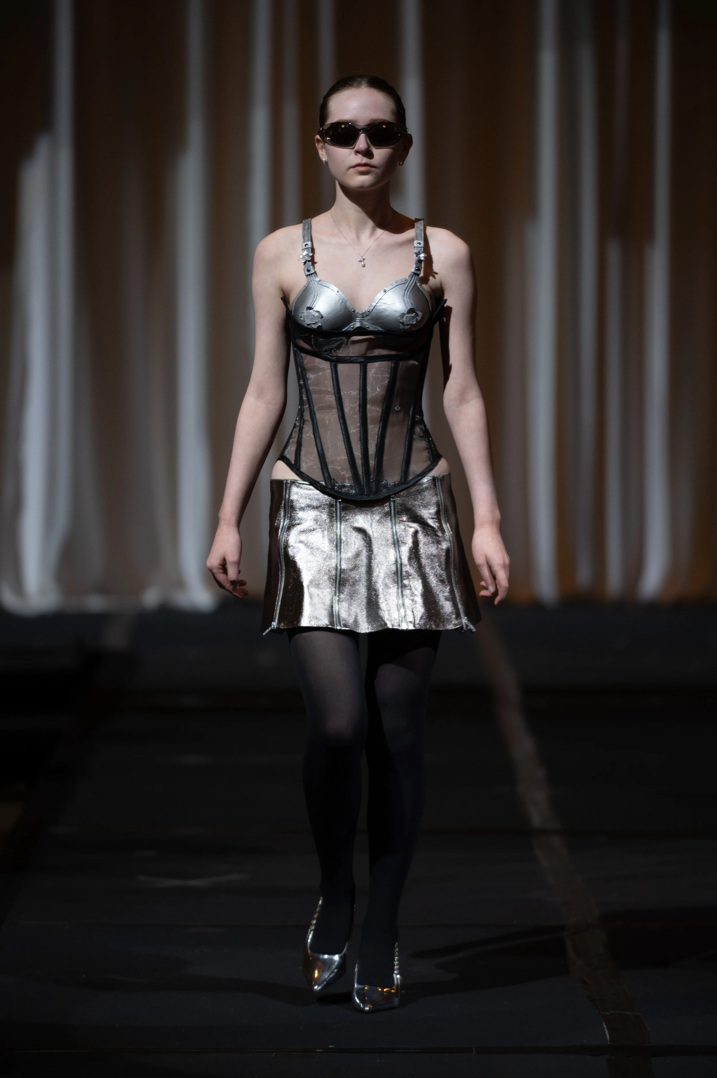 Fashion model walking on runway wearing metallic-top and black skirt with black tights and silver high heels, dark hair tied back, sunglasses, in a dark setting with curtains in the background.