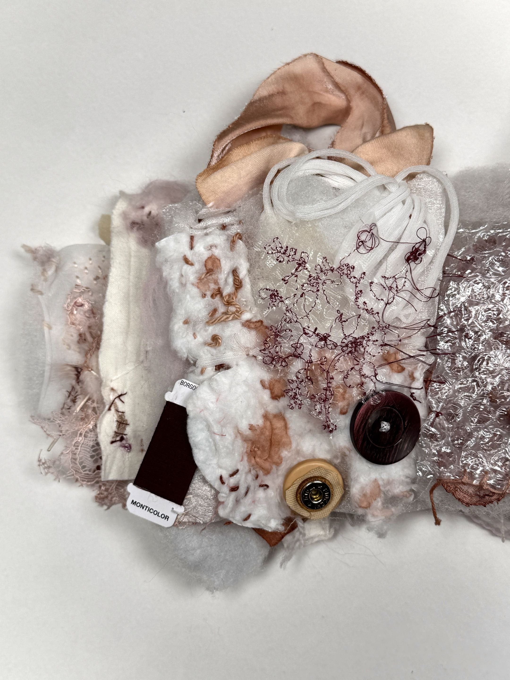 Various fabric scraps, buttons, and sewing supplies arranged on a flat surface, including lace, satin, embroidered fabrics, and thread, some labeled with tags.
