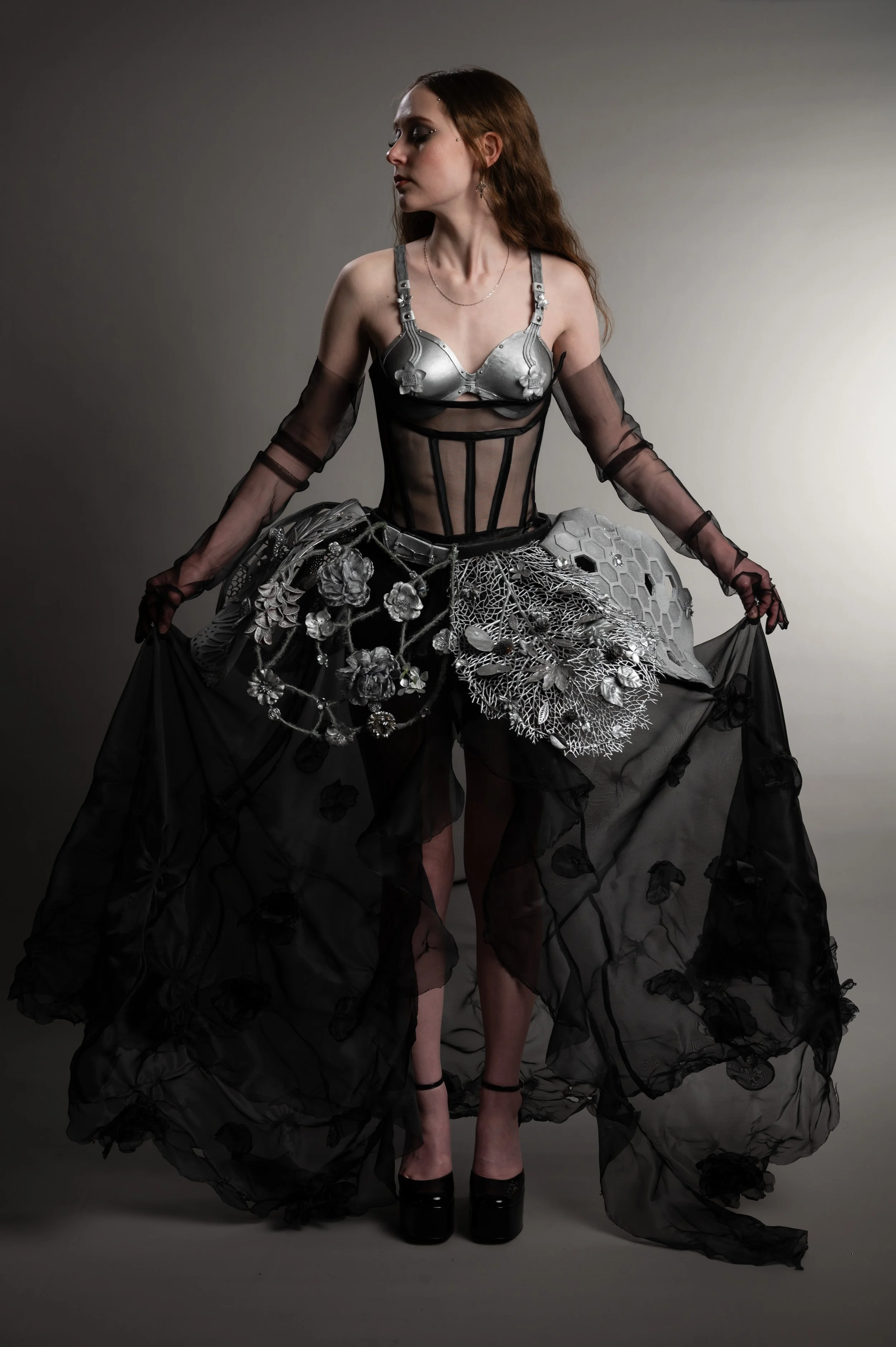 A woman wearing a sheer black dress with floral embellishments and high heels, holding the dress out to reveal the intricate design and her legs, standing against a plain backdrop.