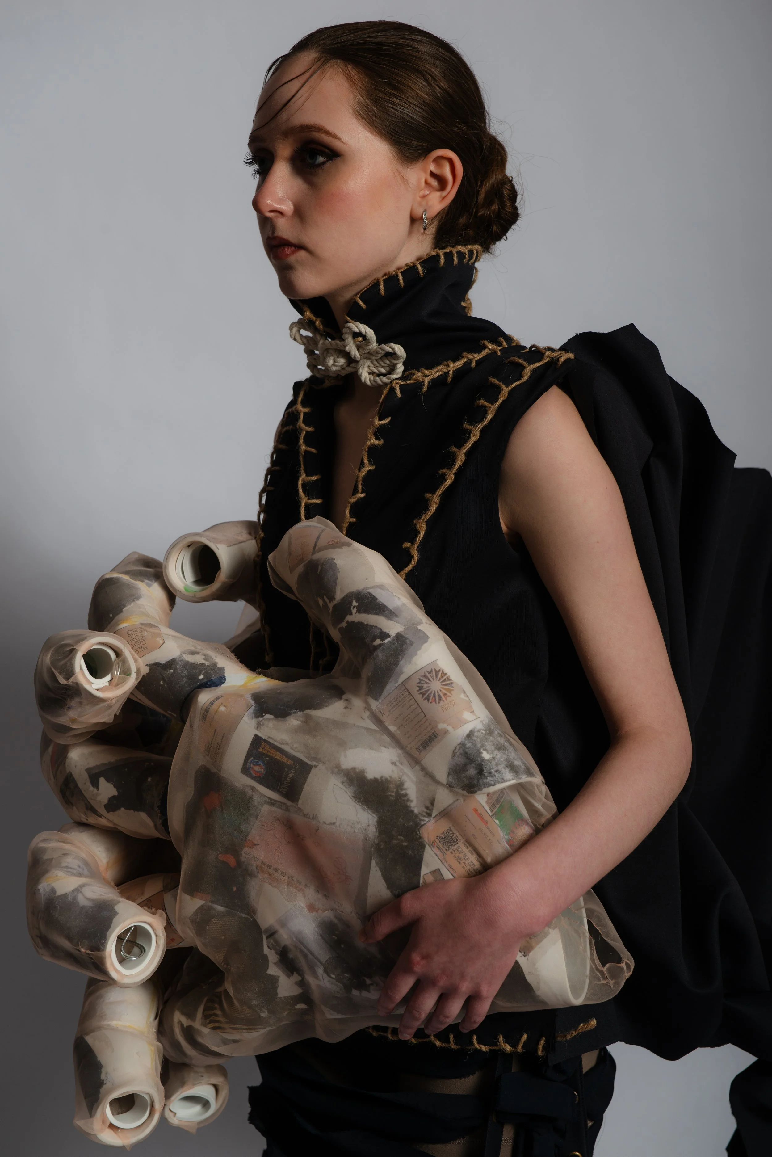 A woman with dark hair styled back, wearing a black outfit with gold-stitch detailing, holds a large, sculptural, hand-shaped object made of collage-like material featuring black and white images, standing against a plain light gray background.