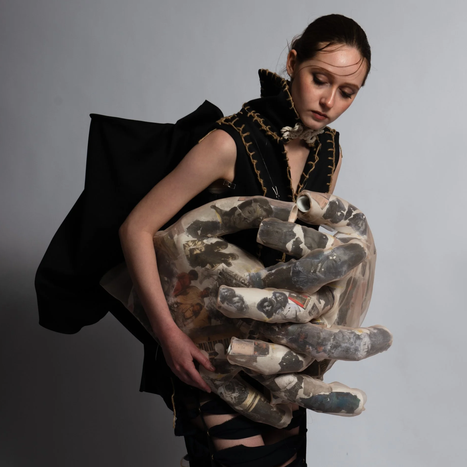 A model dressed in a dark, collared jacket with gold stitching, holds a large sculptural form of two interlocking hands, made of rubbings from old photographs.