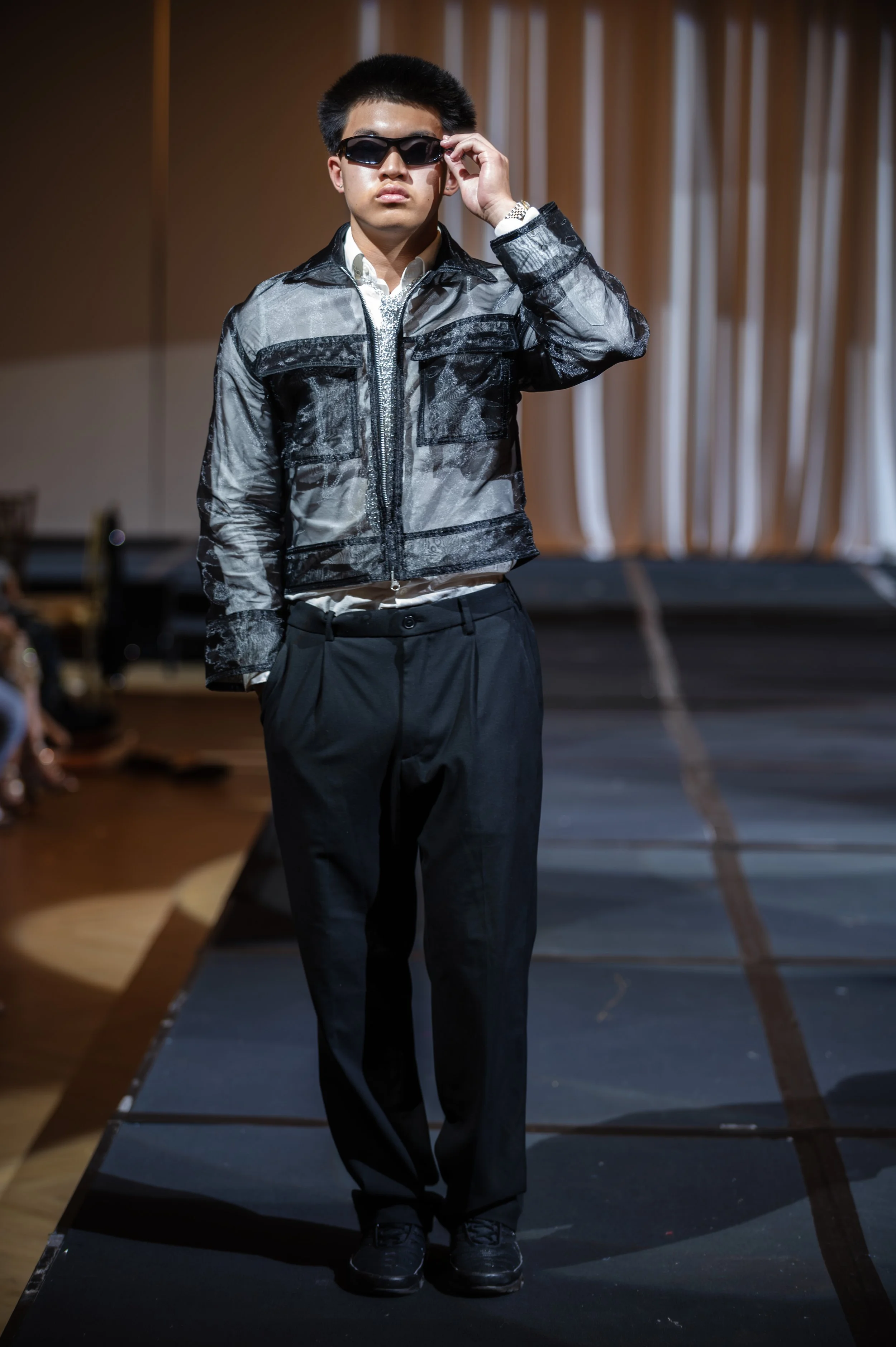 Young man wearing a silver and black patterned jacket, black pants, and black shoes, standing on a runway with curtains in the background, adjusting his sunglasses.