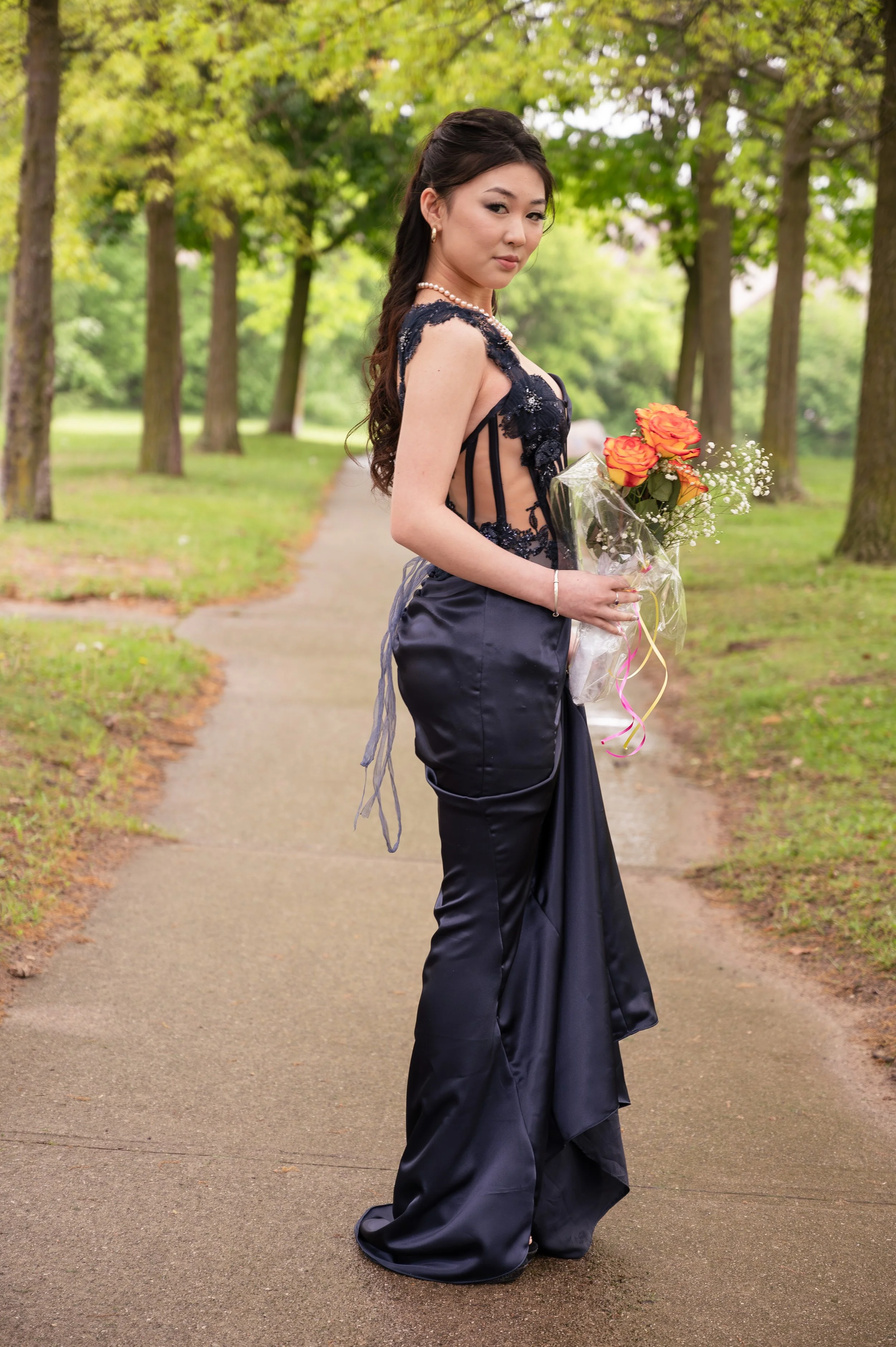 A woman in an elegant black gown holding a bouquet of orange roses stands on a park path surrounded by green trees.