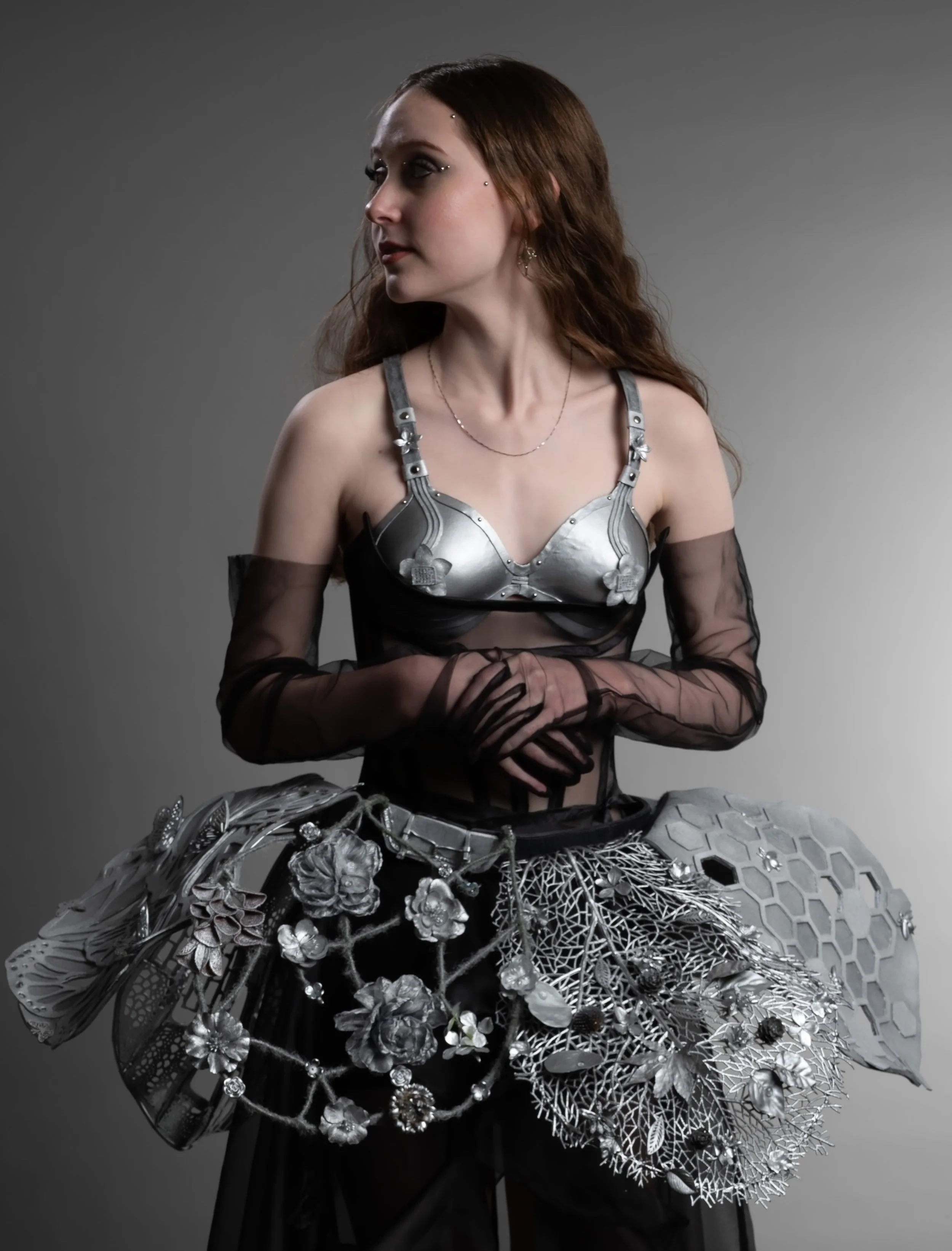 A model dressed in futuristic metallic and black attire with floral embellishments, standing against a gray background.