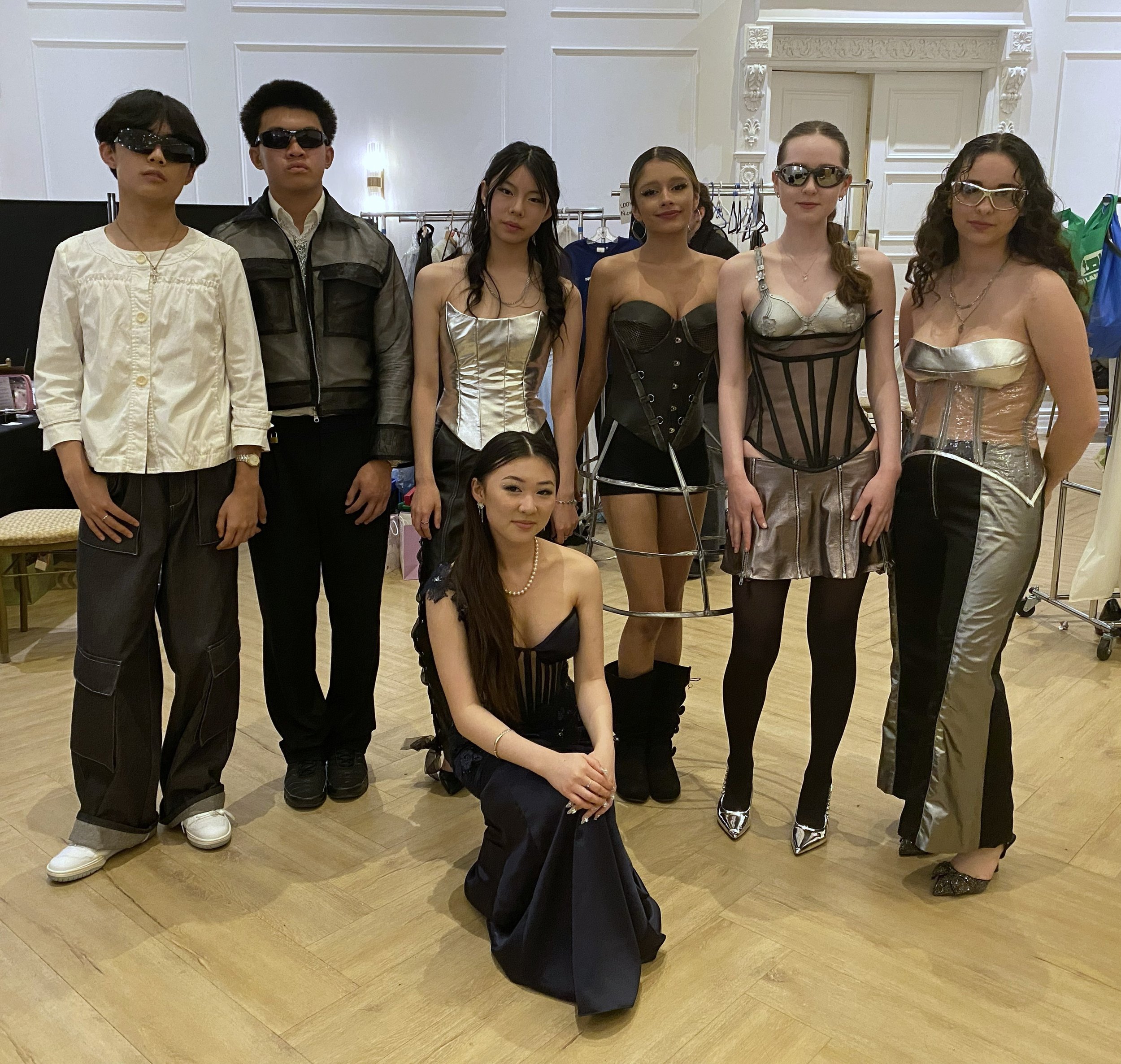 A group of seven young people, six standing and one sitting in front, posing for a photo in a room with white decorative walls. The individuals are dressed in stylish clothing, some wearing glasses or sunglasses, with a clothing rack visible in the background.