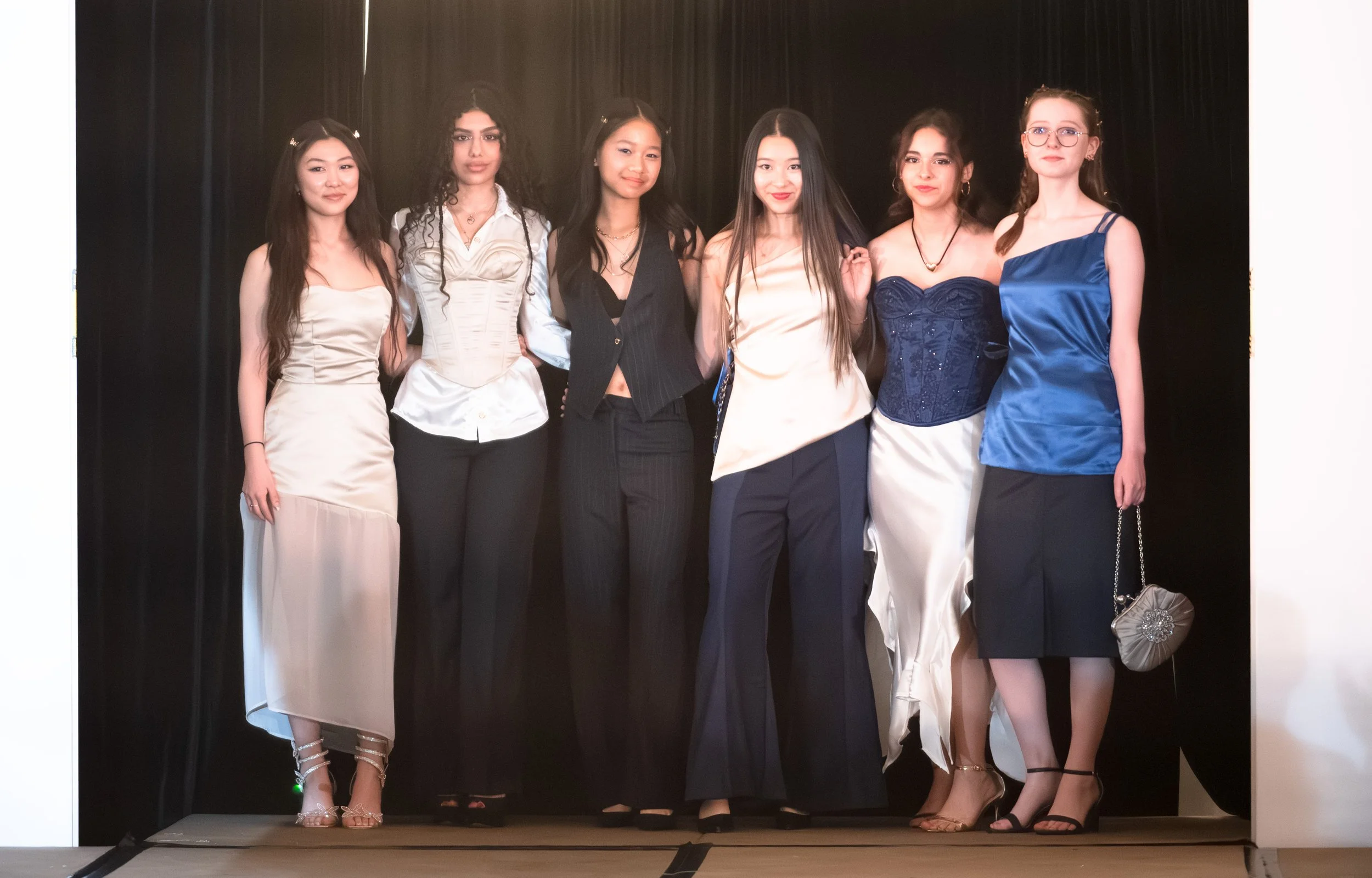 Six women standing together on a stage with a black curtain backdrop, dressed in formal and semi-formal attire.