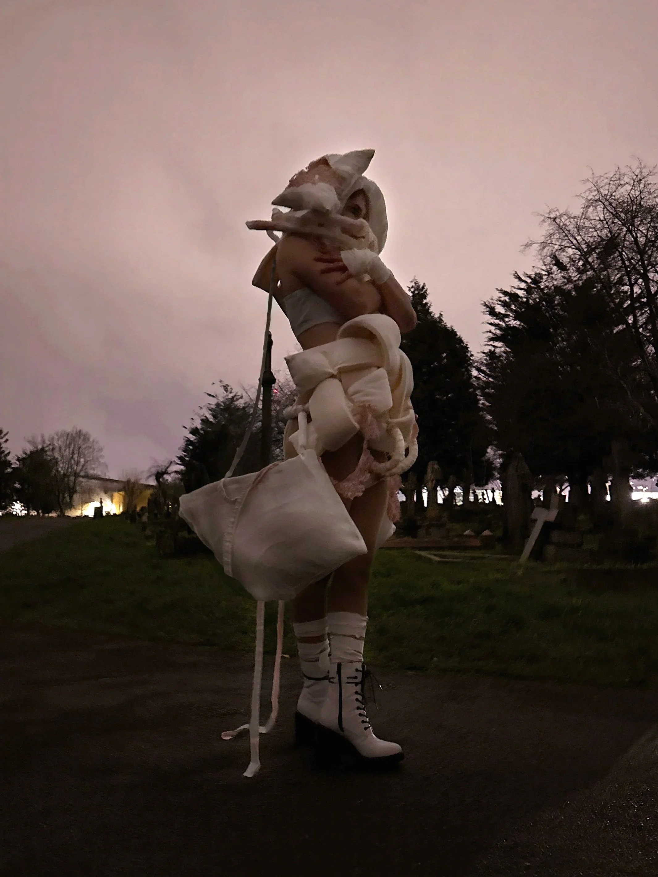 A person wearing white avant-garde clothing, including high-heeled boots, gloves,, standing outside at dusk with trees and park benches in the background.