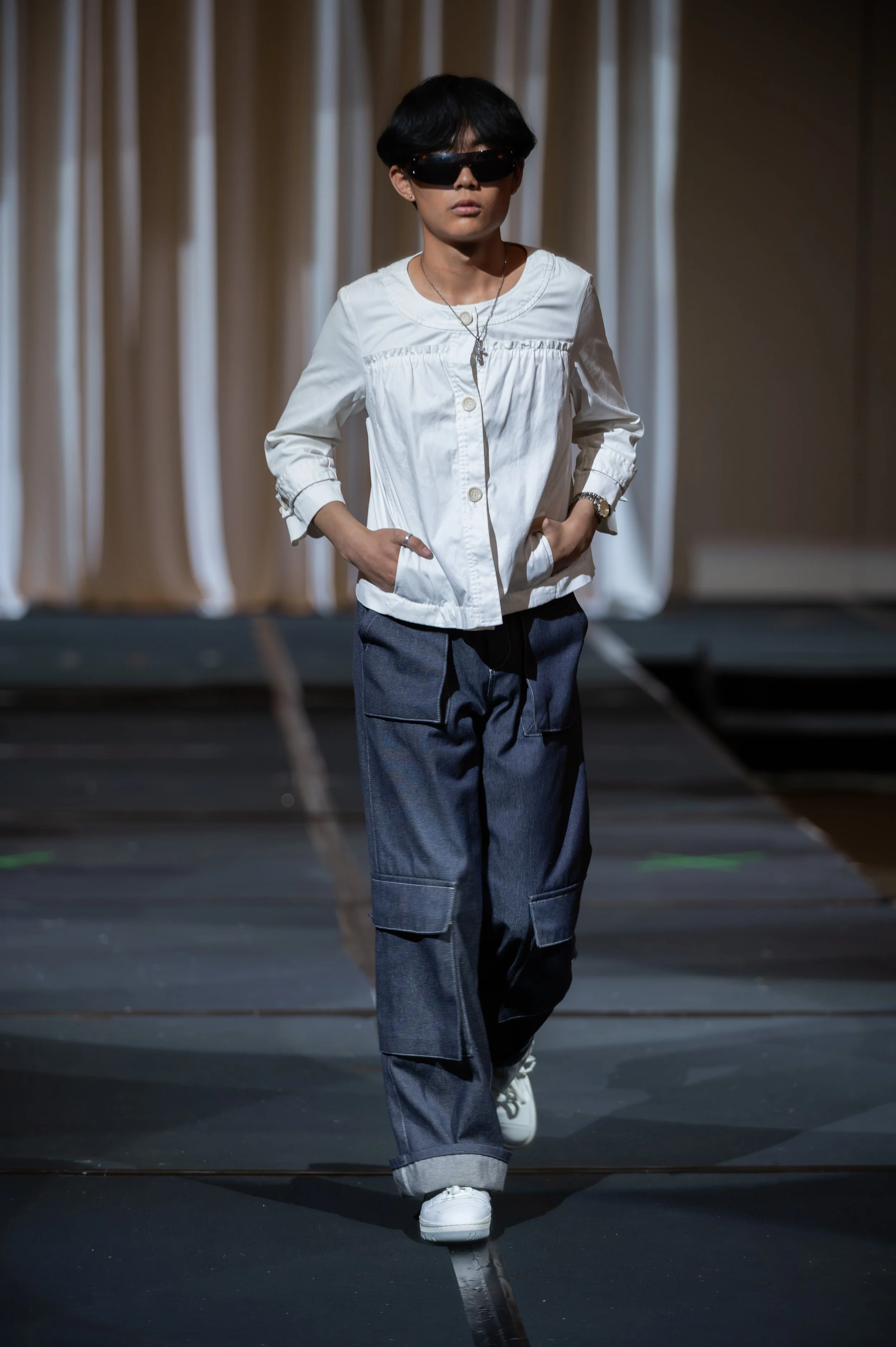 A person walking on a runway with hands in pockets, wearing sunglasses, a white buttoned shirt, loose denim jeans, and white sneakers.