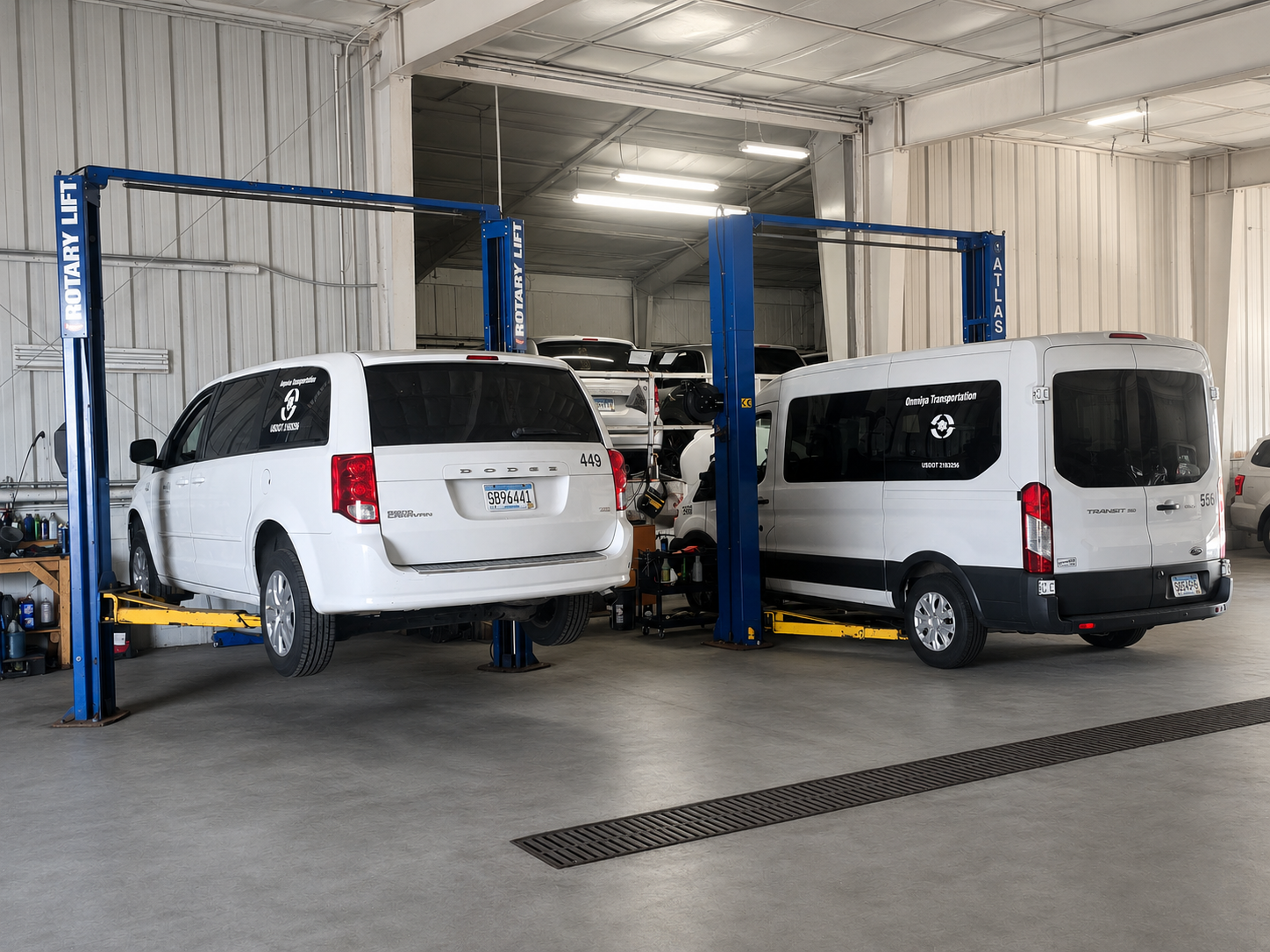 Indoor auto repair shop with two white vehicles on hydraulic lifts, one being a Dodge minivan and the other a Ford Transit van, surrounded by tools and equipment.