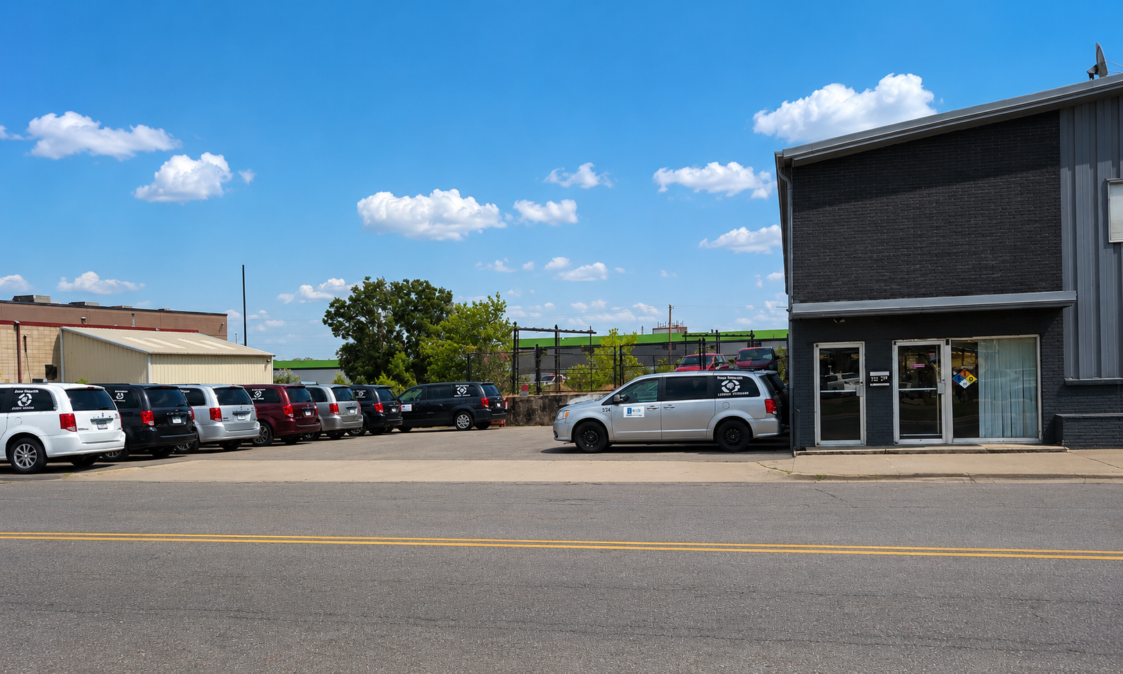A row of parked cars in front of a black brick building on a sunny day with a blue sky and some clouds.