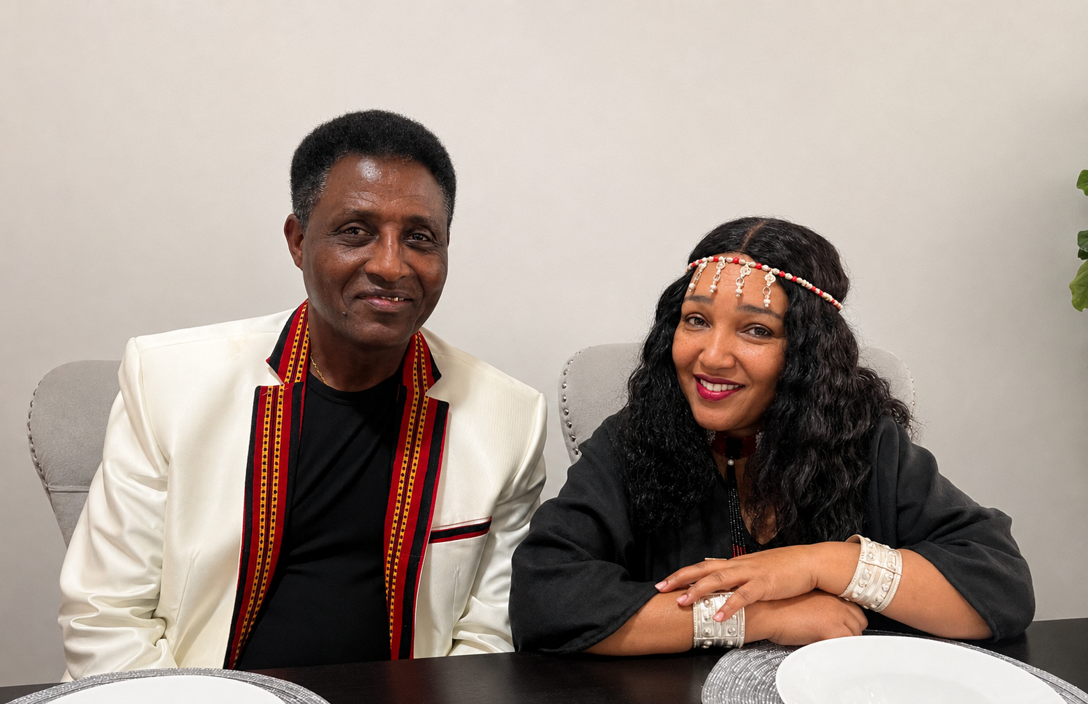 A man and woman sitting at a table, smiling, dressed in party or cultural attire, with a plain background.