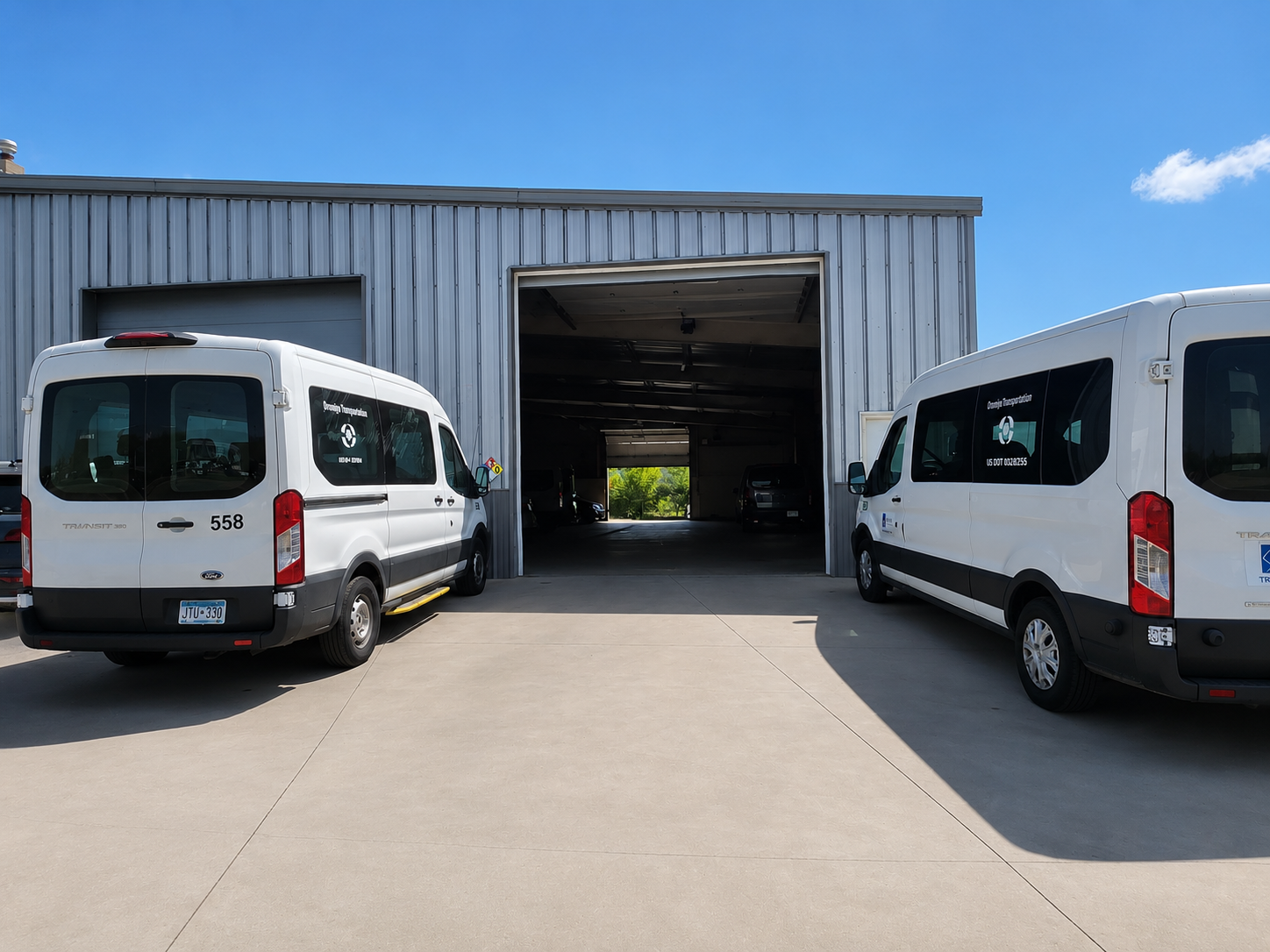 Two white transit vans parked outside a large metal building with an open garage door, revealing a dark interior and cars inside. The sky is blue with a few clouds.
