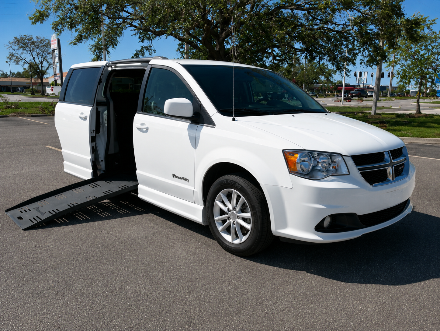 White mobility van with a side door open, equipped with a ramp for wheelchair access, parked in an empty lot with trees and a shopping area in the background.