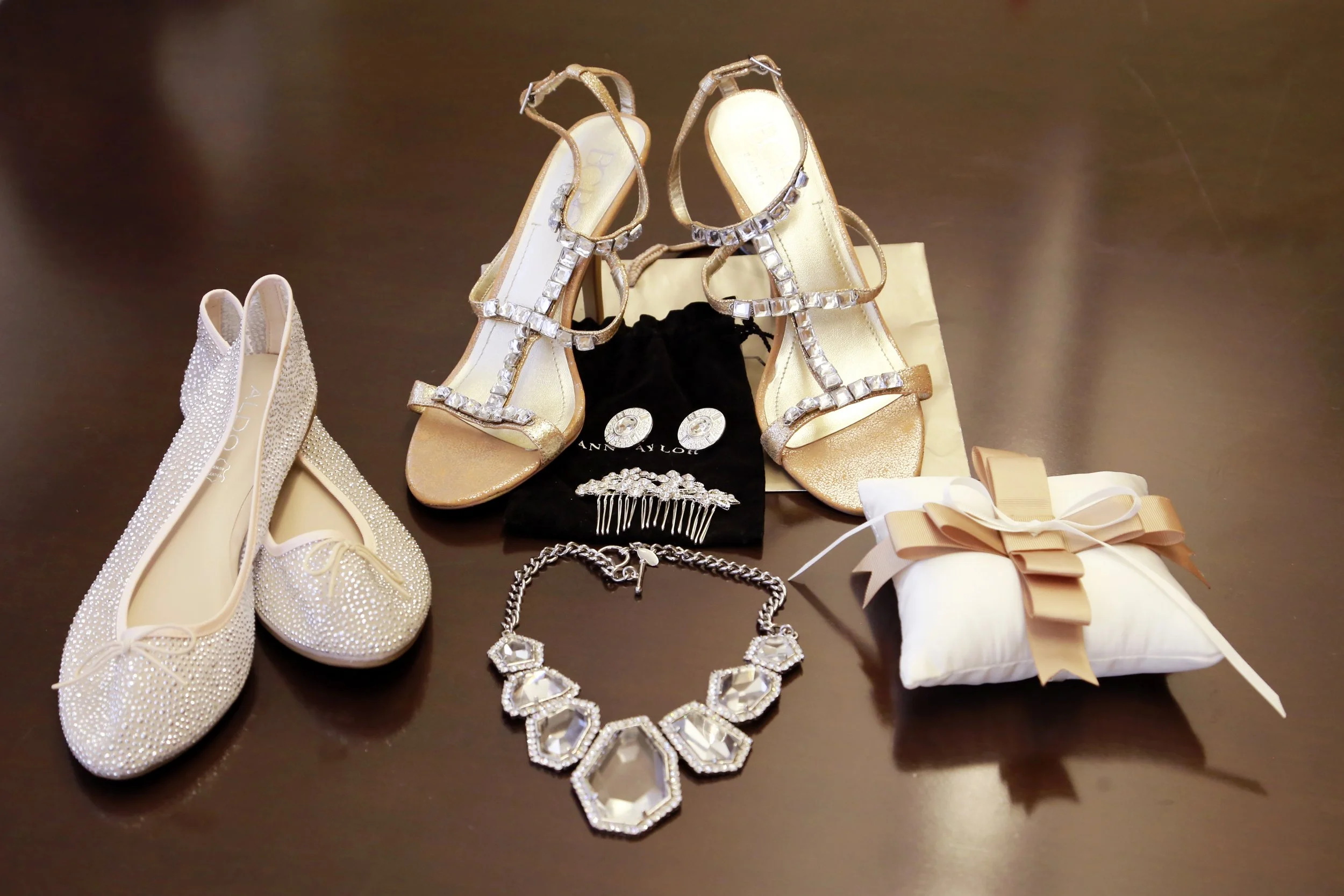 A collection of jewelry and shoes, including a pair of white shoes covered in rhinestones, a pair of gold high-heeled sandals with rhinestone embellishments, a pair of earrings, a bracelet, and a wrapped gift with a ribbon, displayed on a dark surface.