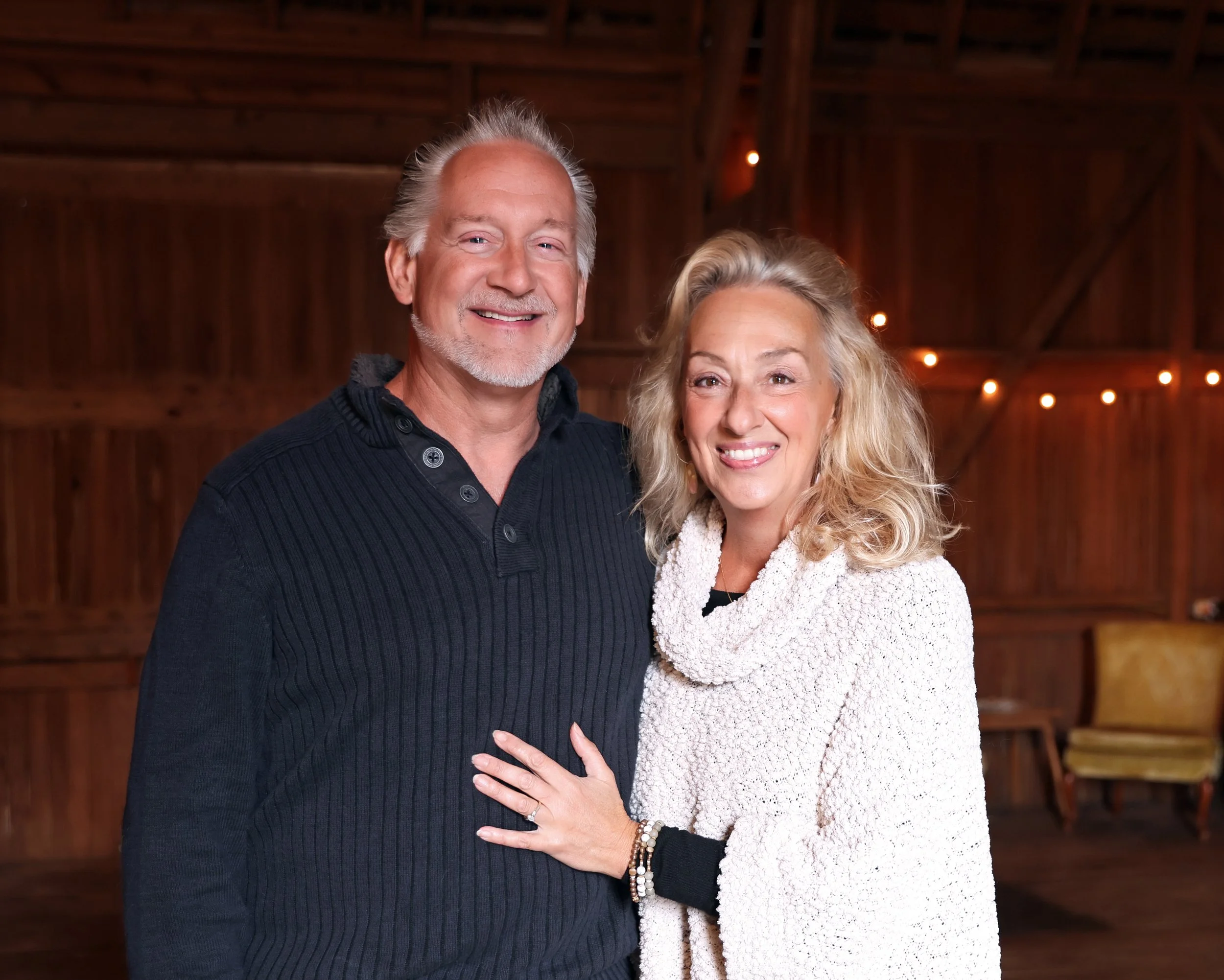 A smiling man with gray hair and a beard wearing a black sweater and a smiling woman with blonde curly hair wearing a white textured sweater, standing together in a wooden barn or rustic setting with string lights in the background.