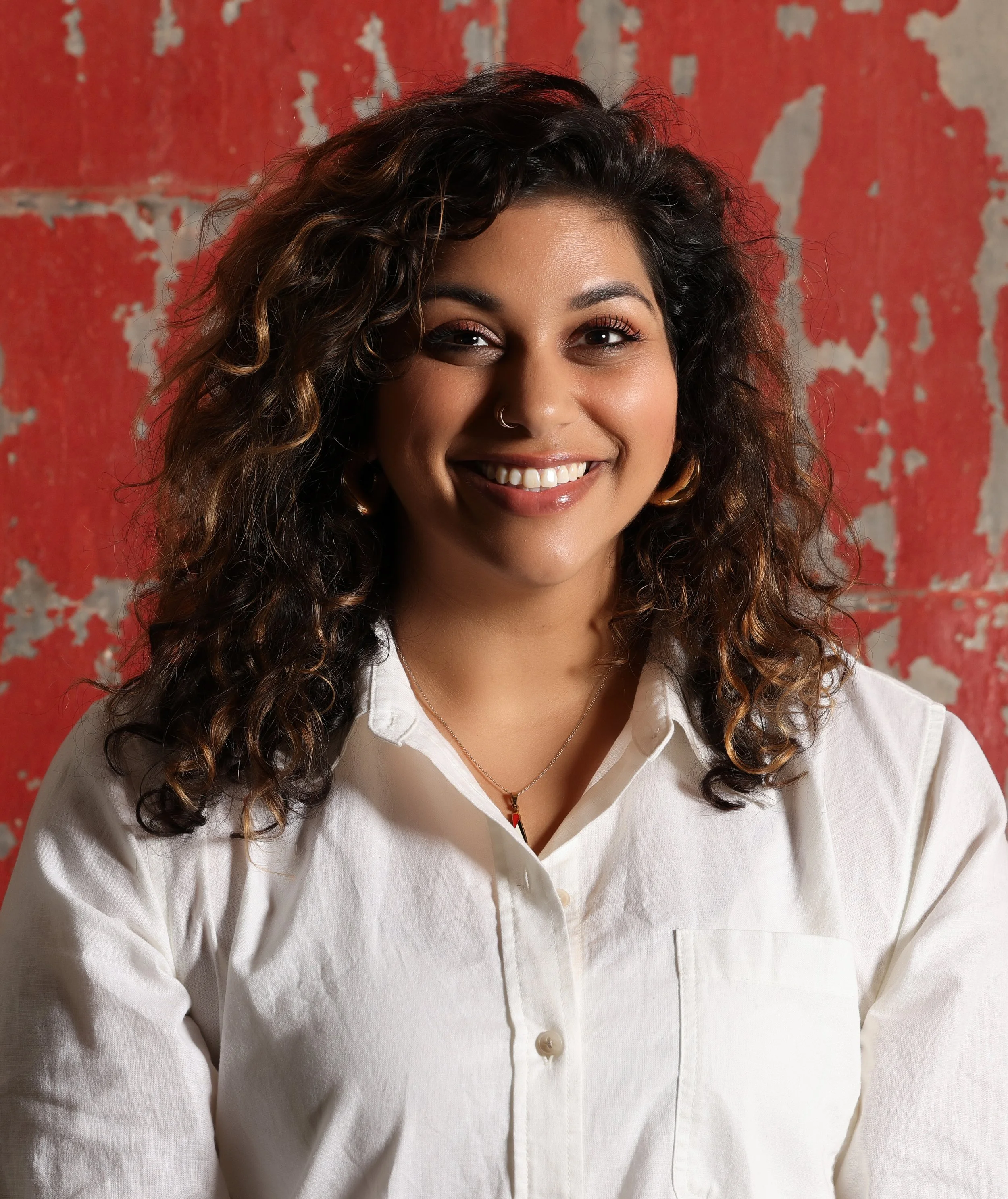 A woman with curly dark brown hair smiling, wearing a white button-up shirt and a necklace with a red pendant. She has a small nose ring in her left nostril. The background features an aged, peeling red wall.