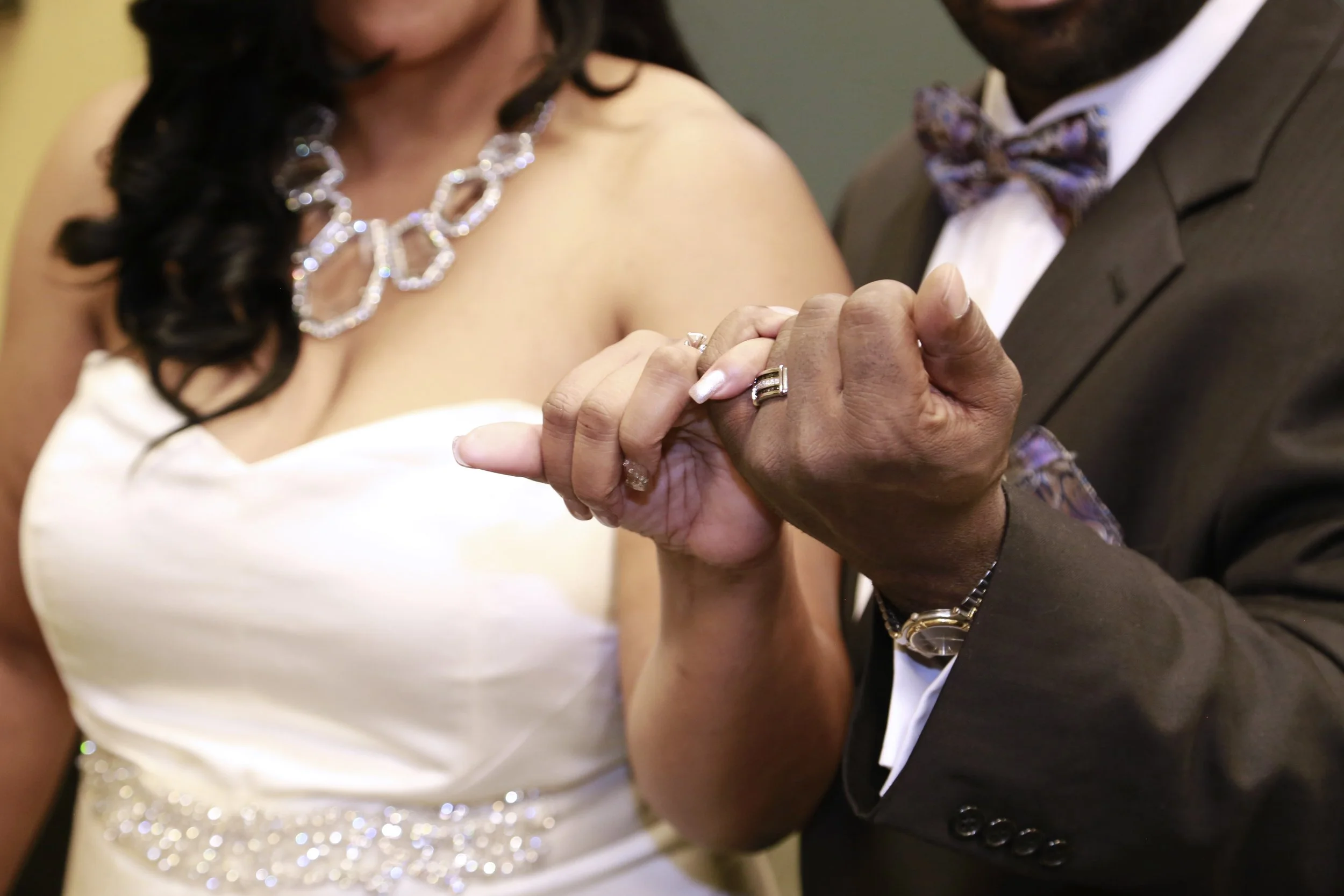 A close-up of a couple holding hands, with wedding rings visible. The woman is wearing a white dress with a sparkling necklace, and the man is dressed in a dark suit with a colorful bow tie.