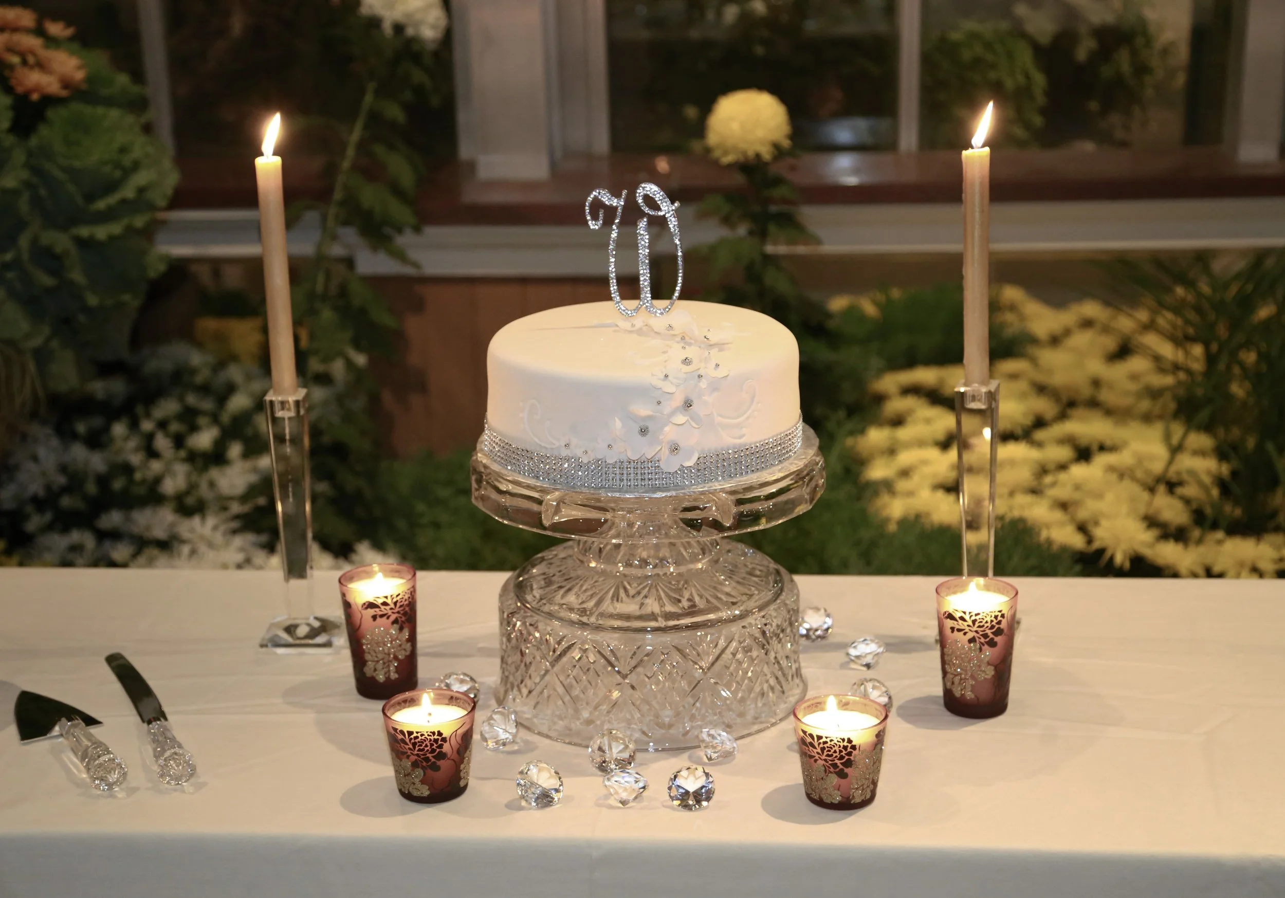 A white birthday cake with silver decorative accents, topped with a glittery '70' glitter cake topper, is set on a glass stand. Surrounding the cake are five lit candles in pink holders with floral patterns, all placed on a white table. The background shows plants and a window.