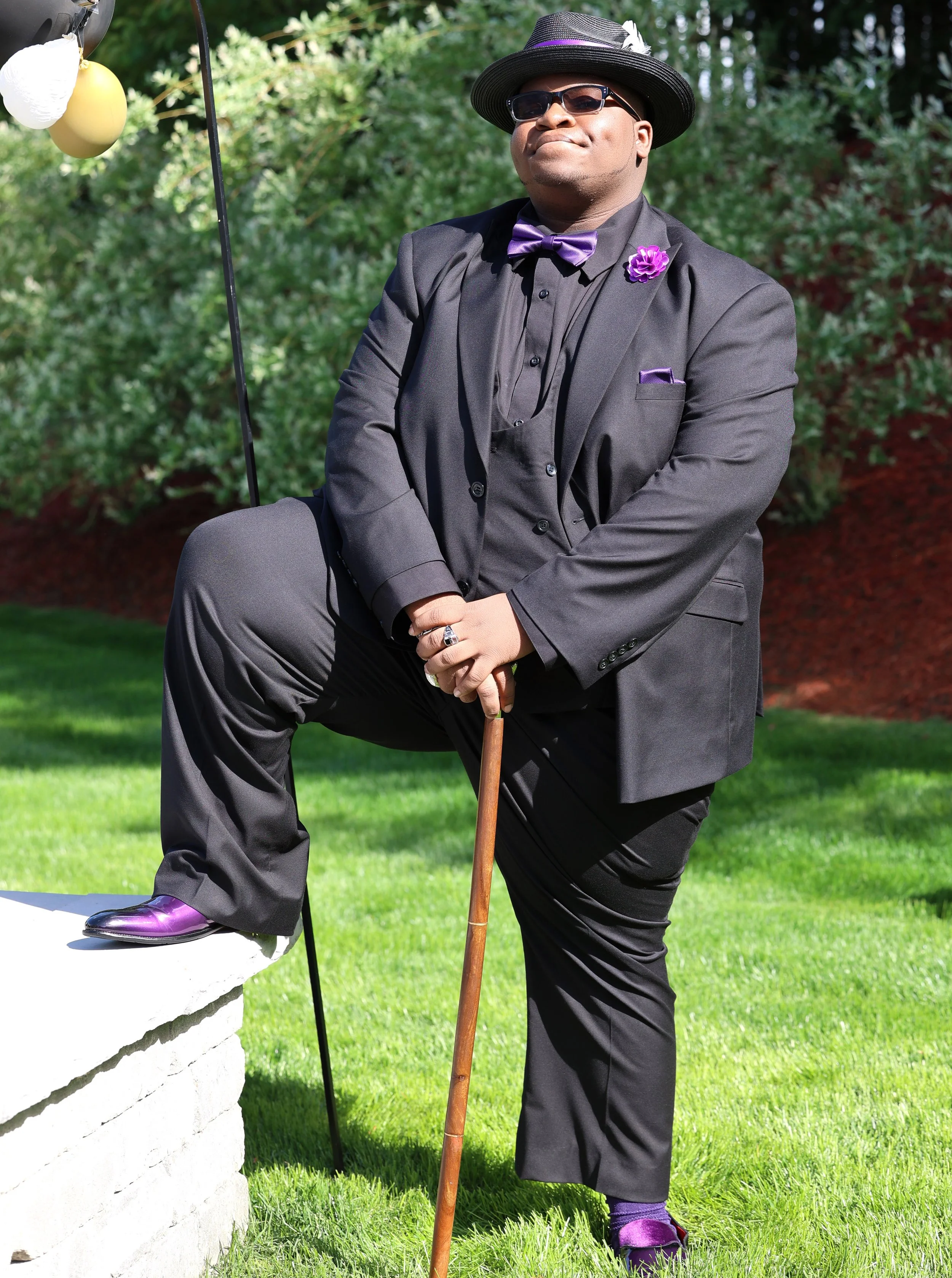 A man dressed in a black suit with purple accessories and a top hat, standing outdoors with one foot on a white brick ledge and holding a walking cane. He is wearing sunglasses and poses confidently against a background of green grass and foliage.