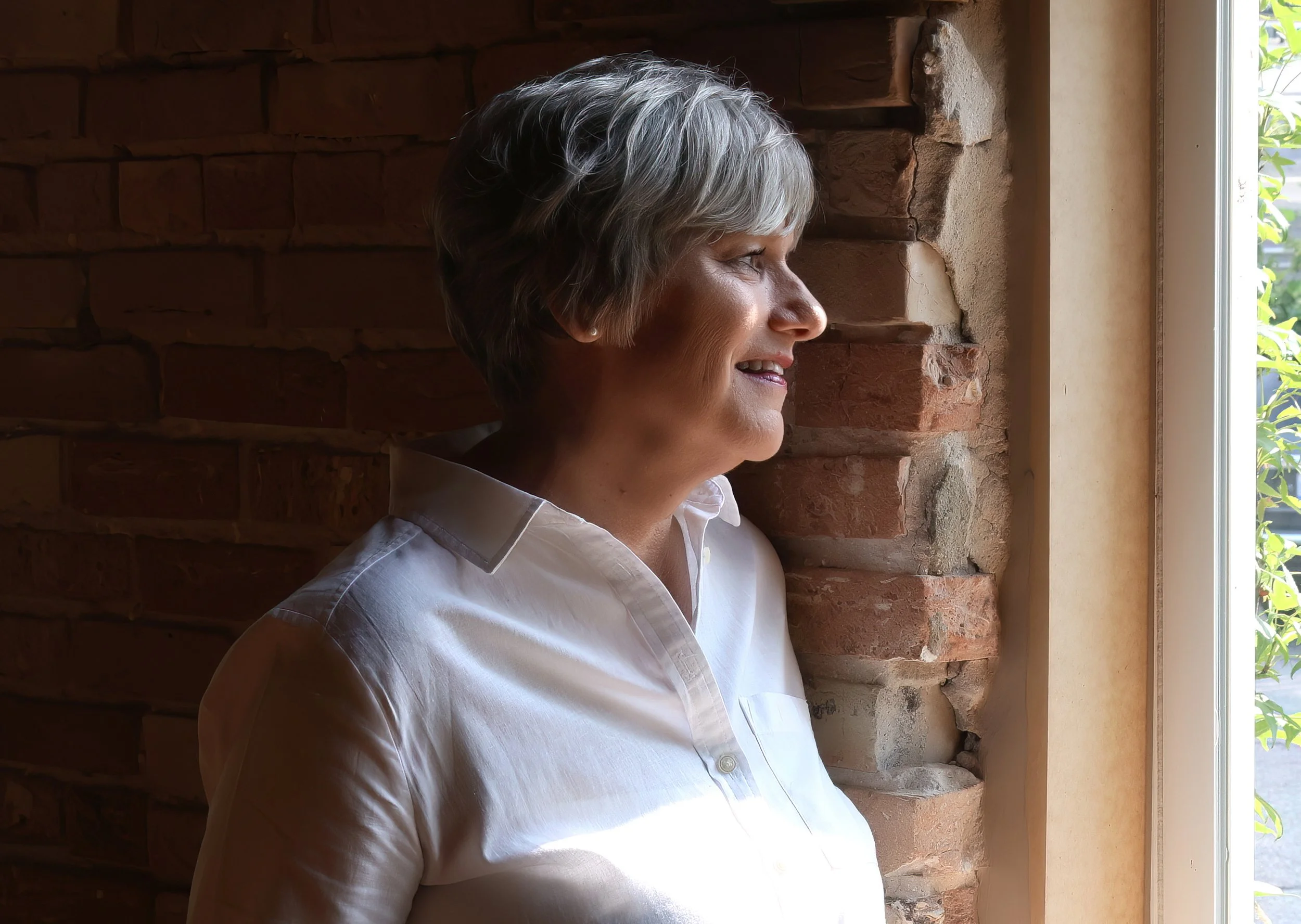 A woman with short gray hair smiling, sitting indoors next to a brick wall with a window showing green foliage outside.