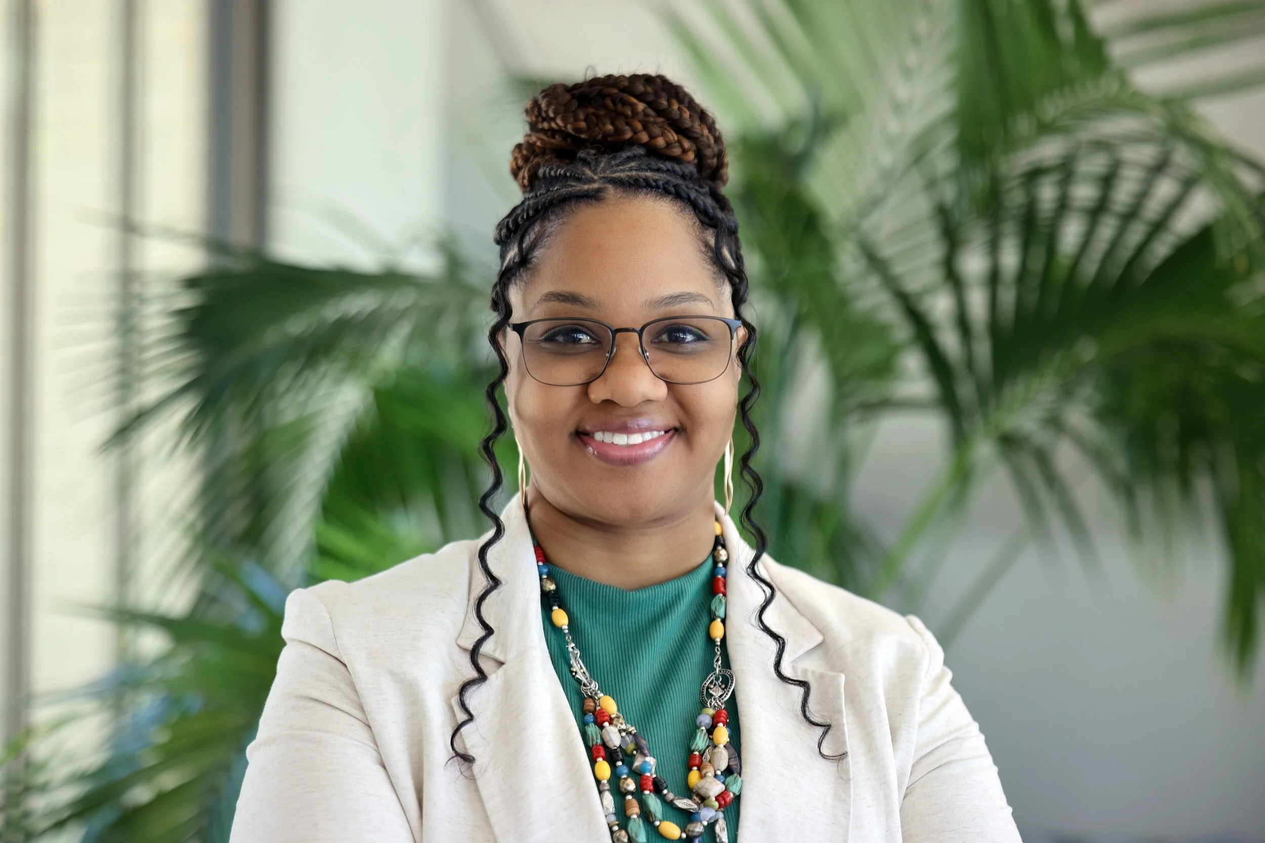 A woman with dark curly hair styled in a large braided bun on top of her head, wearing glasses, a colorful beaded necklace, and a light blazer, smiling in front of green tropical plants.