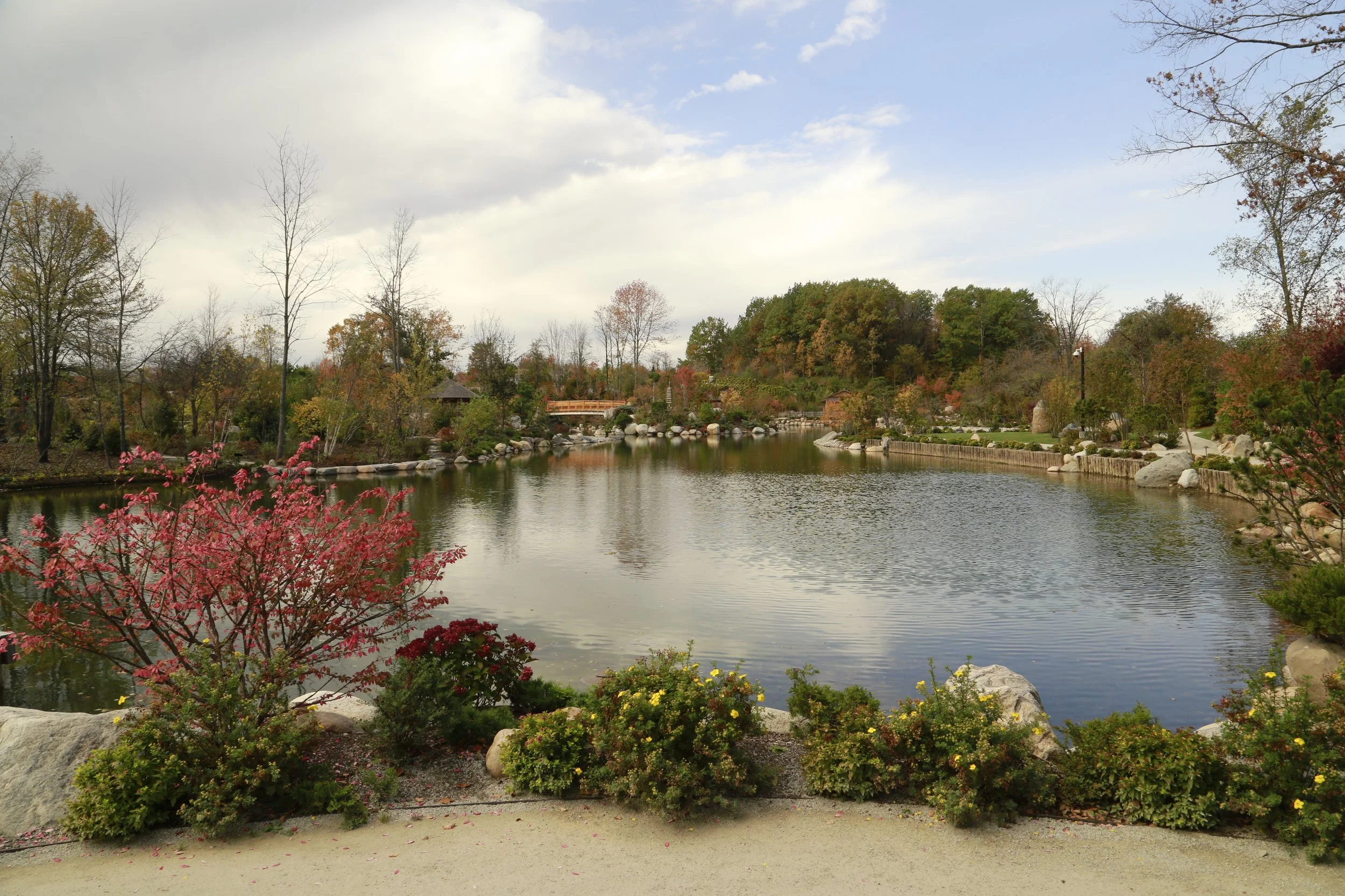 A scenic park with a pond, trees displaying fall foliage, a bridge, and a cloudy sky.